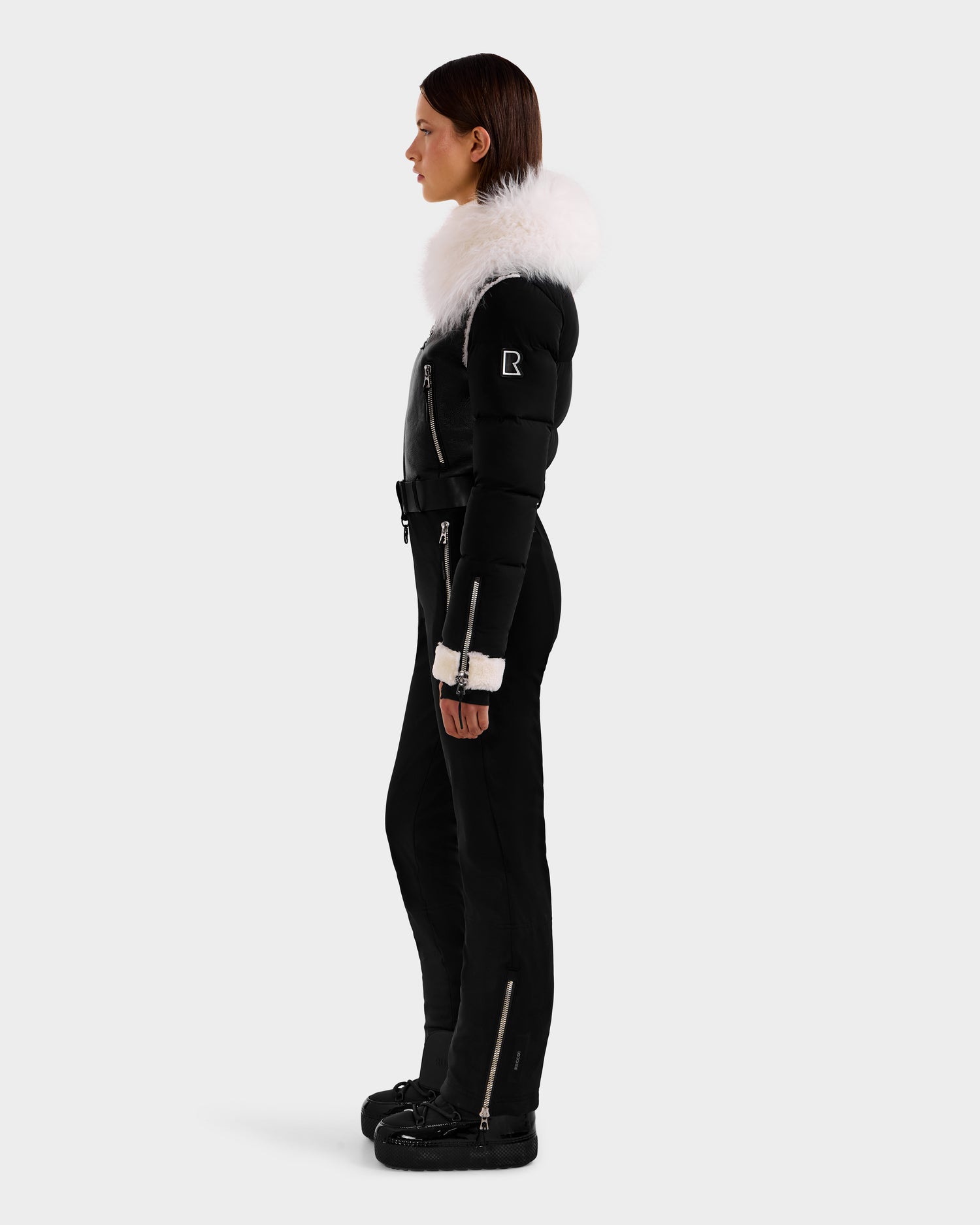 A woman in profile models the CHYNTIA - 8125611 Black ski suit by Rudsak, featuring a black fitted design, white fur-lined hood, cream cuffs, and black boots against a plain light gray background.