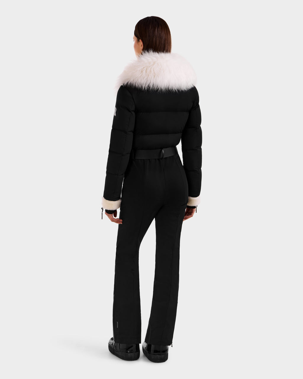 A woman faces away in the Rudsak CHYNTIA - 8125611 Black puffer jacket, featuring a wide white fur collar and cuffs, paired with black belted pants and boots against a light background.