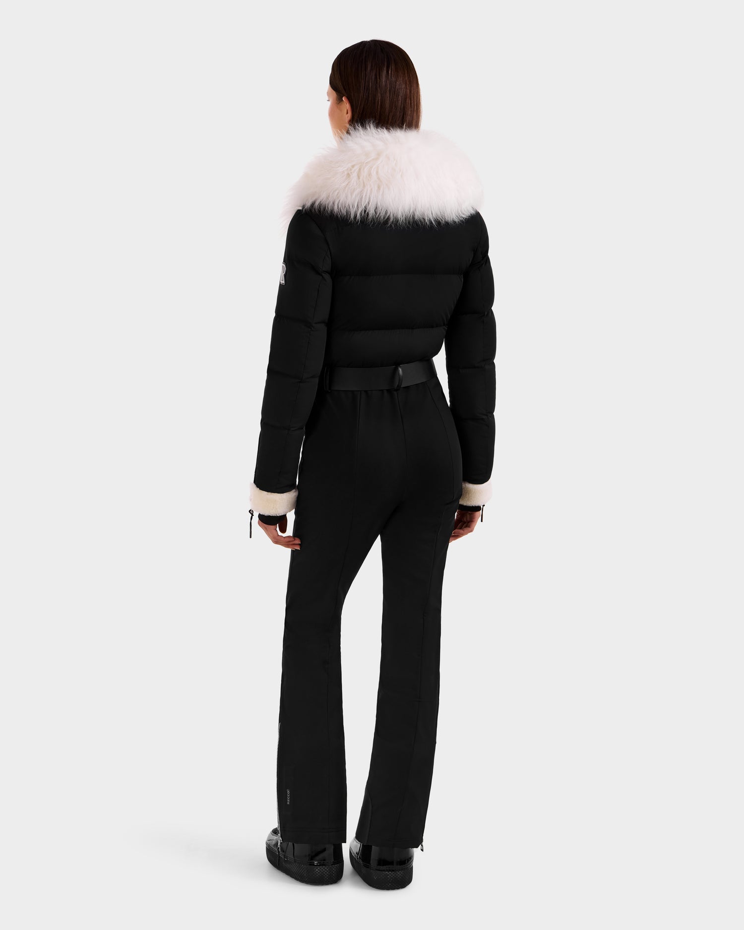 A woman faces away in the Rudsak CHYNTIA - 8125611 Black puffer jacket, featuring a wide white fur collar and cuffs, paired with black belted pants and boots against a light background.