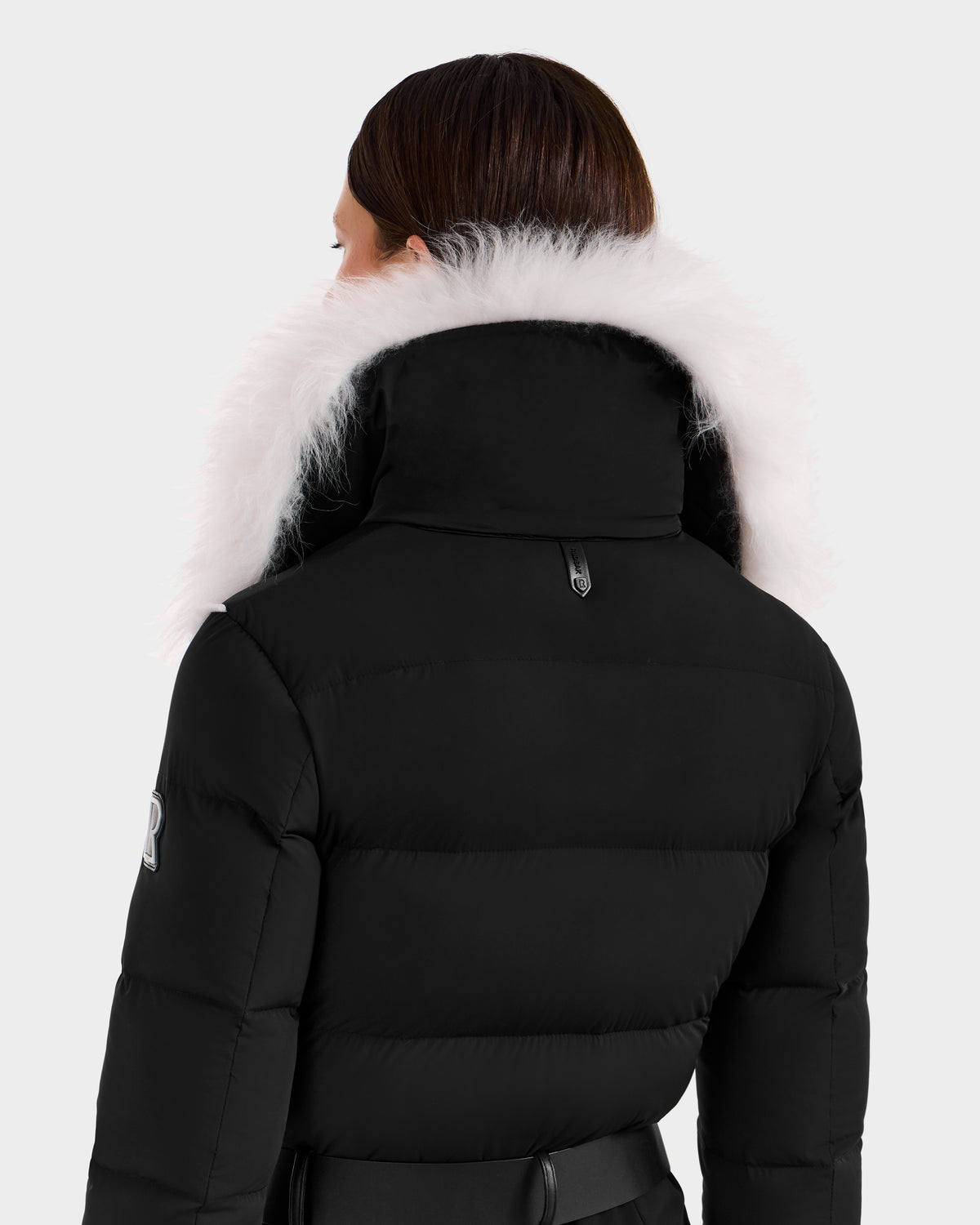 A person with brown hair, seen from behind, wears the Rudsak CHYNTIA - 8125611 Black puffer jacket featuring a high collar with fluffy white fur trim, set against a plain, light background.