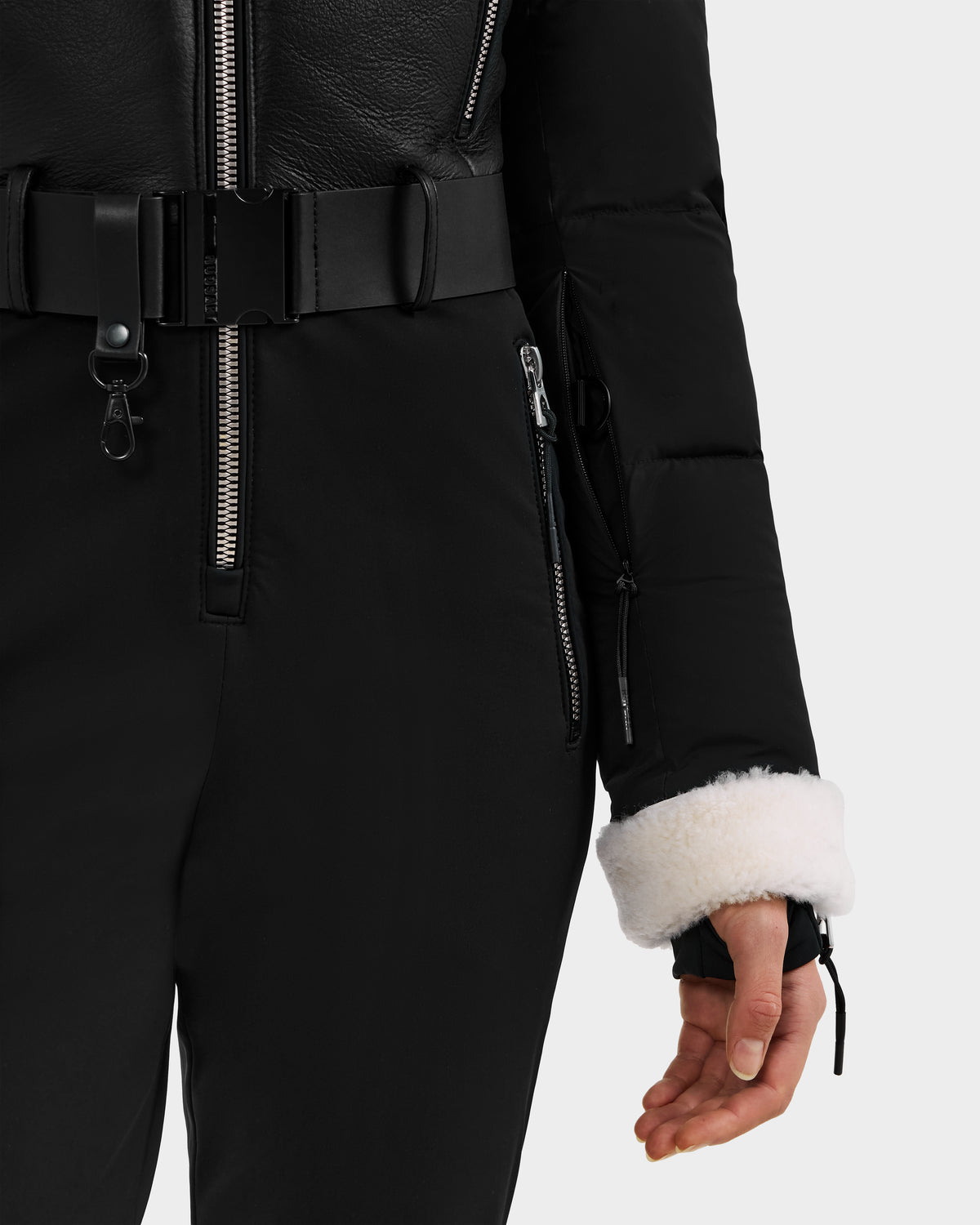 A person wearing the Rudsak CHYNTIA - 8125611 Black women’s ski suit with a belted waist, black pants, and white faux fur cuffs stands with one arm slightly extended against a plain background.