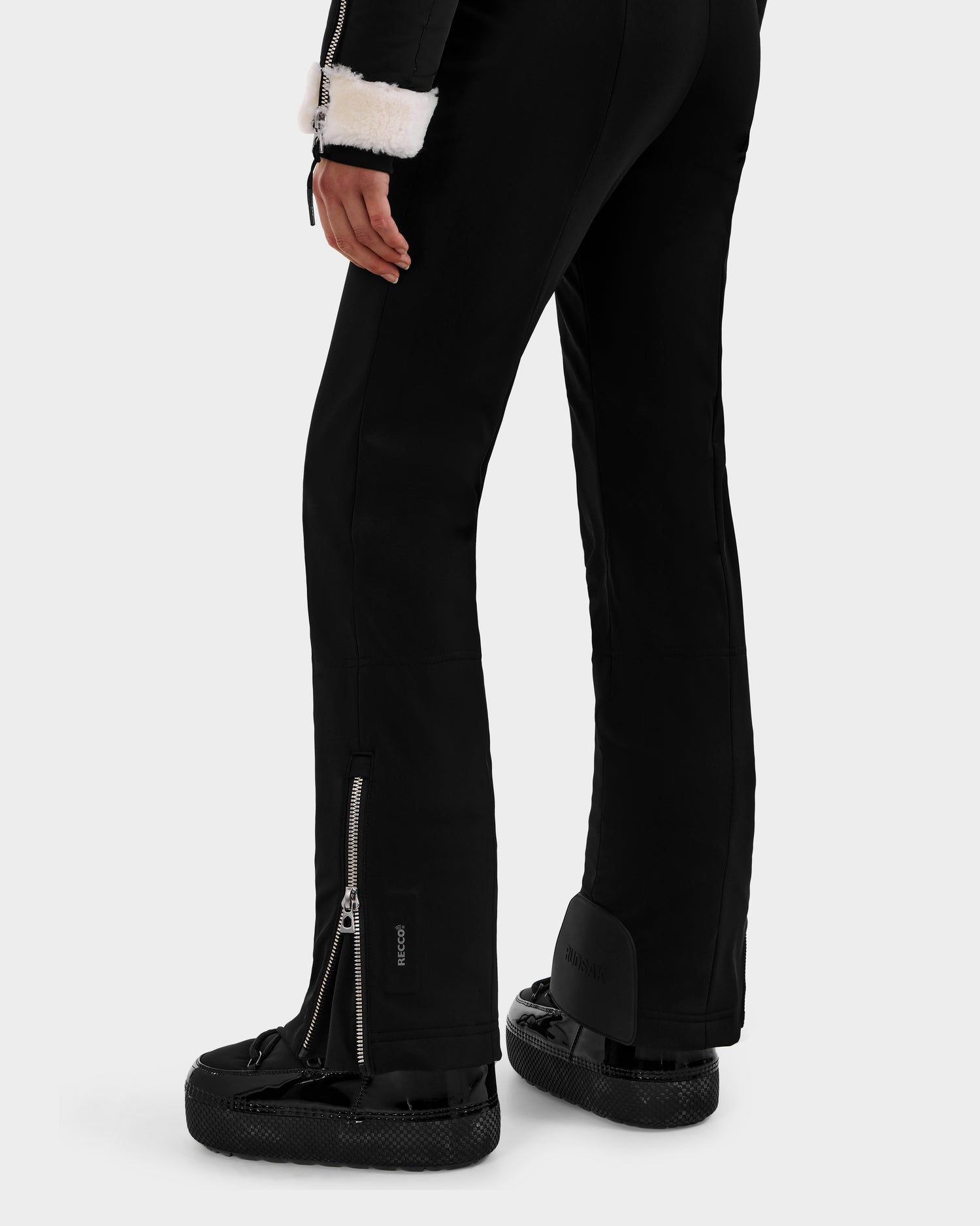 A person models the Rudsak CHYNTIA - 8125611 Black pants, featuring back leg zippers, styled with black platform shoes and a white faux fur-trimmed sleeve for a chic ski-inspired look against a plain background.
