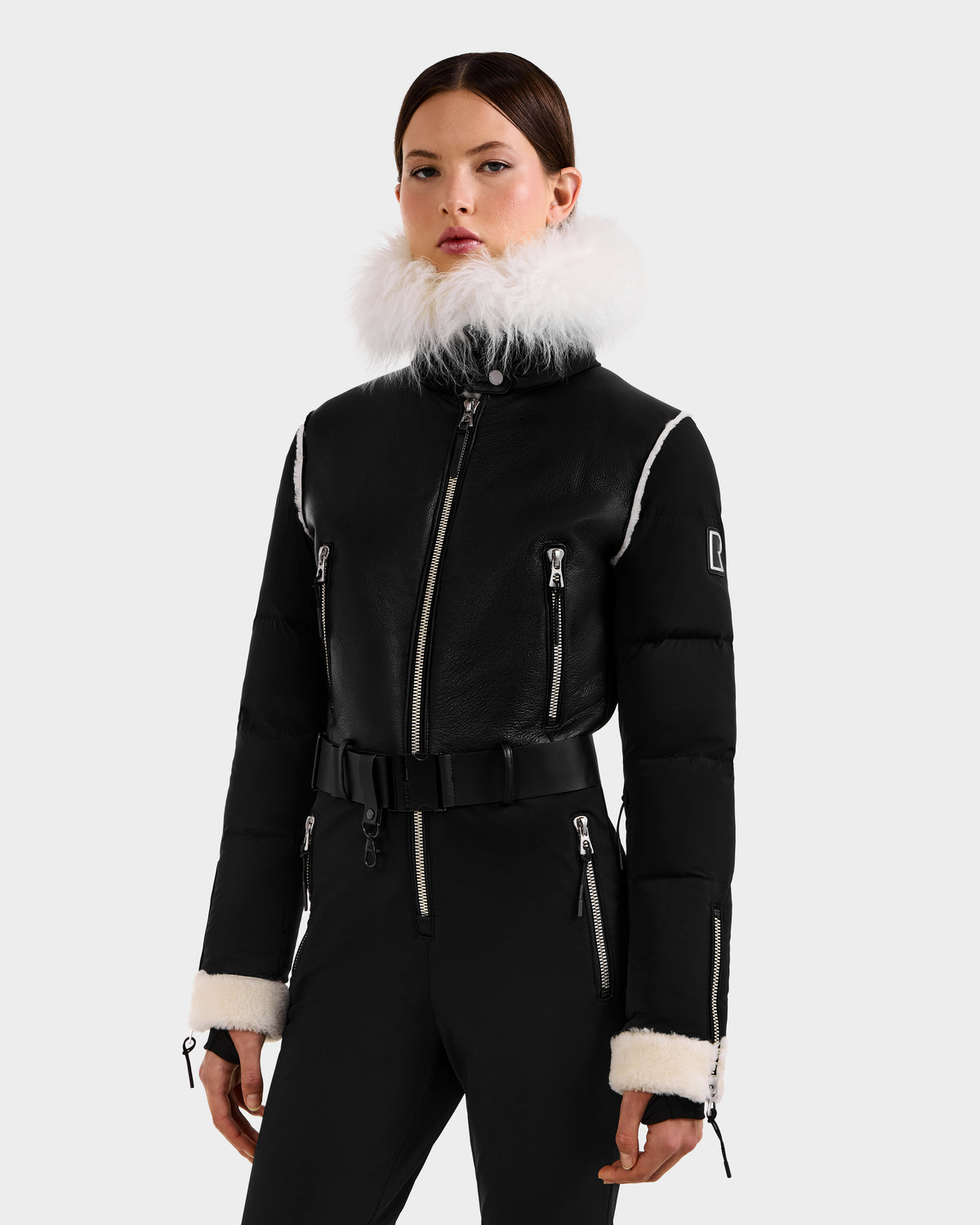 A woman models the Rudsak CHYNTIA - 8125611 Black jacket, featuring white fur trim, silver zippers, and a waist belt—capturing the modern elegance of a high-fashion winter wardrobe.