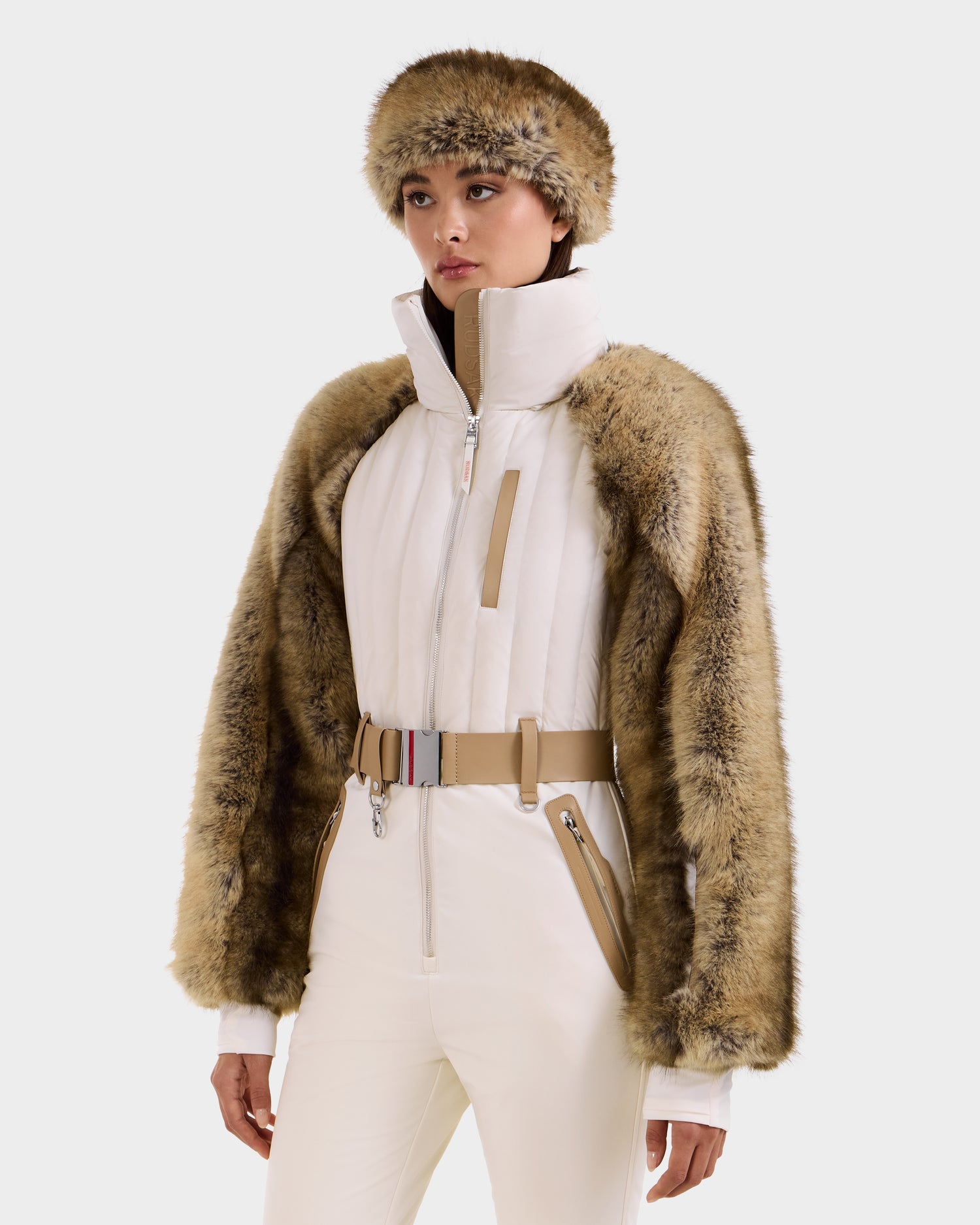 A woman wears the Rudsak DESTINY - 8124727 Vanilla ski suit, a white one-piece with a high collar, wide beige belt, and tan faux fur trim on sleeves and shoulders, paired with a matching faux fur hat, against a plain white background.
