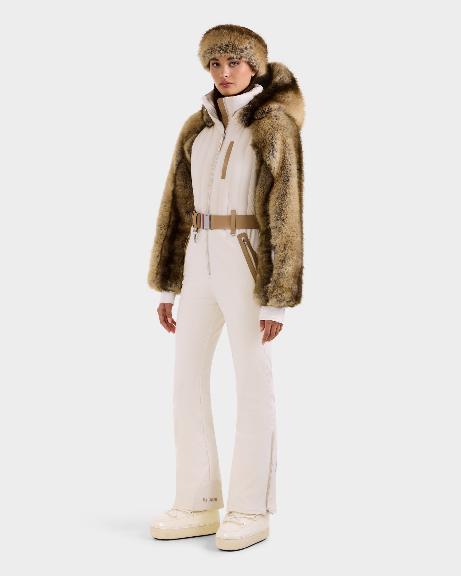 A person wears the Rudsak DESTINY - 8124727 Vanilla ski suit with beige boots, a wide belt, a brown faux fur jacket over their shoulders, and a matching fur hat, standing against a plain light background.