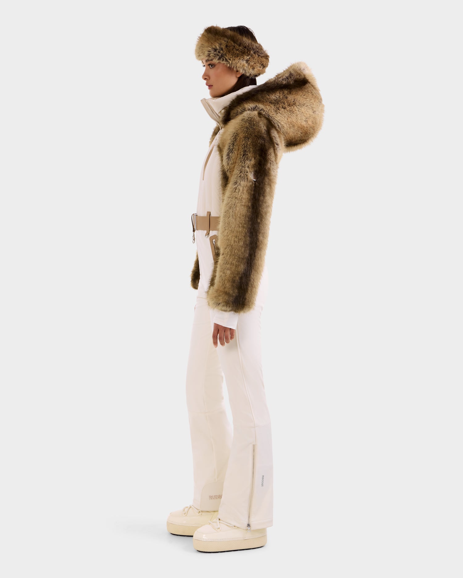 A person stands in profile wearing the Rudsak DESTINY - 8124727 Vanilla faux fur hooded jacket and matching fur headband, cream ski suit pants, and white boots against a plain white background.