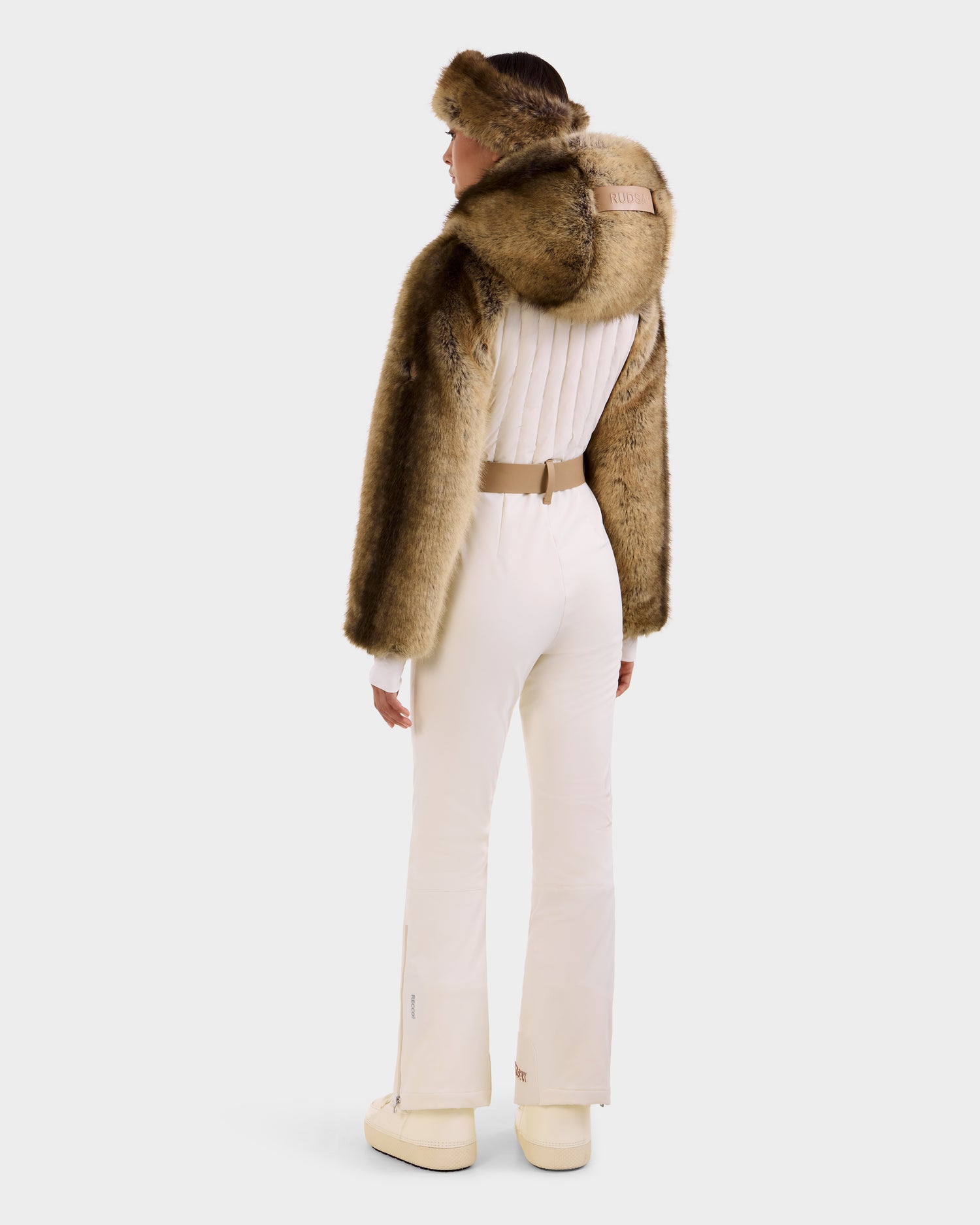 A woman faces away in the Rudsak DESTINY - 8124727 Vanilla faux fur hooded cape with a matching headband, cream long-sleeve top, belted pants, and boots against a plain light background.
