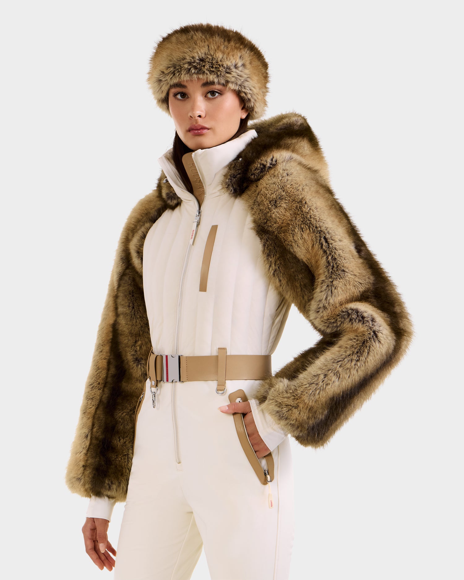 A woman poses confidently in the Rudsak DESTINY - 8124727 Vanilla one-piece ski suit with a belted, hooded jacket and brown faux fur trim, paired with a matching fur hat, against a plain background.