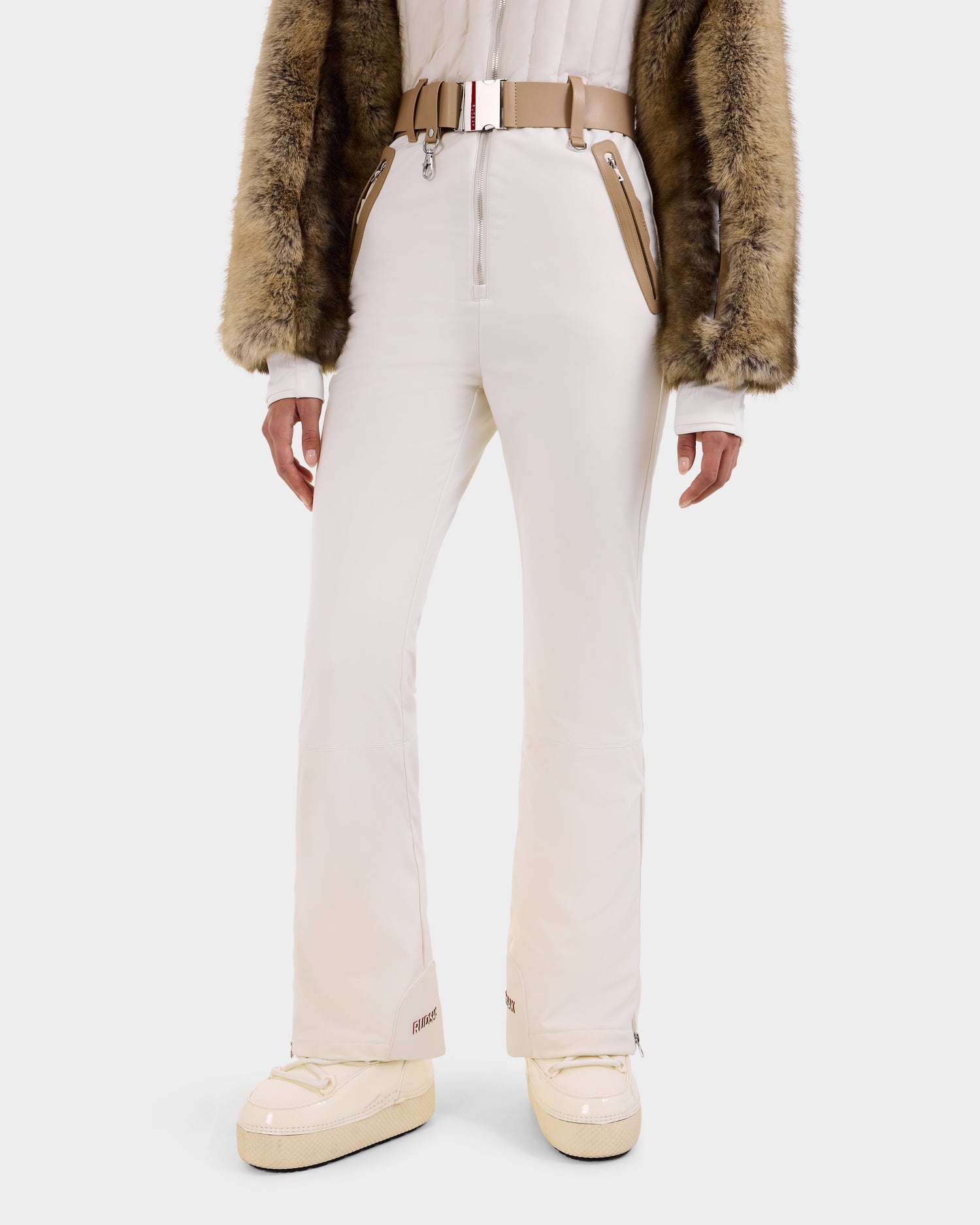 A person wearing Rudsak DESTINY - 8124727 Vanilla women's ski pants with zippered pockets, styled with a beige belt, white top, brown faux fur jacket, and beige boots stands against a white background.