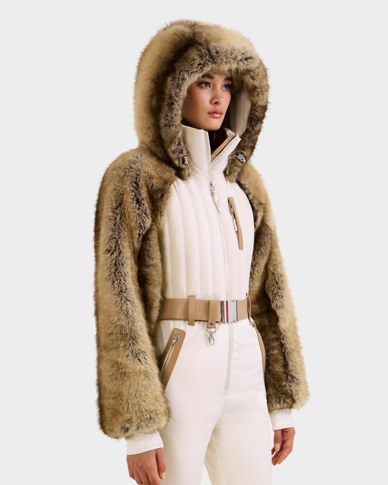 A woman showcases the Rudsak DESTINY - 8124727 Vanilla winter outfit with a tan faux fur hooded jacket, white quilted zip-up top, matching pants, and beige belt—chic inspiration for a stylish women’s ski suit look.