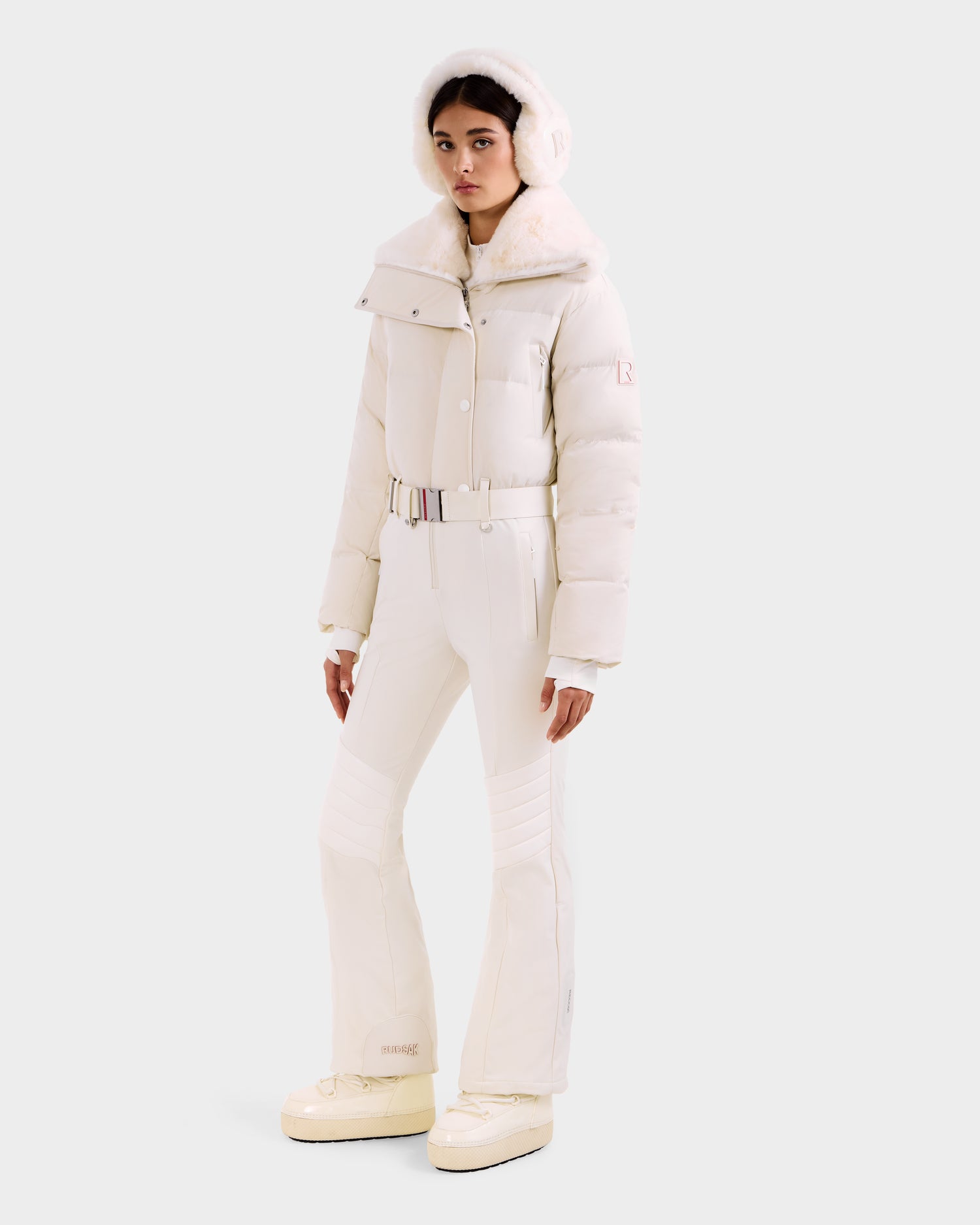 A woman wears the Rudsak LOVE - 8124724 Vanilla off-white ski suit, featuring a wide collar, belted matching pants, and platform boots with earmuffs—a chic winter look perfect for the slopes.