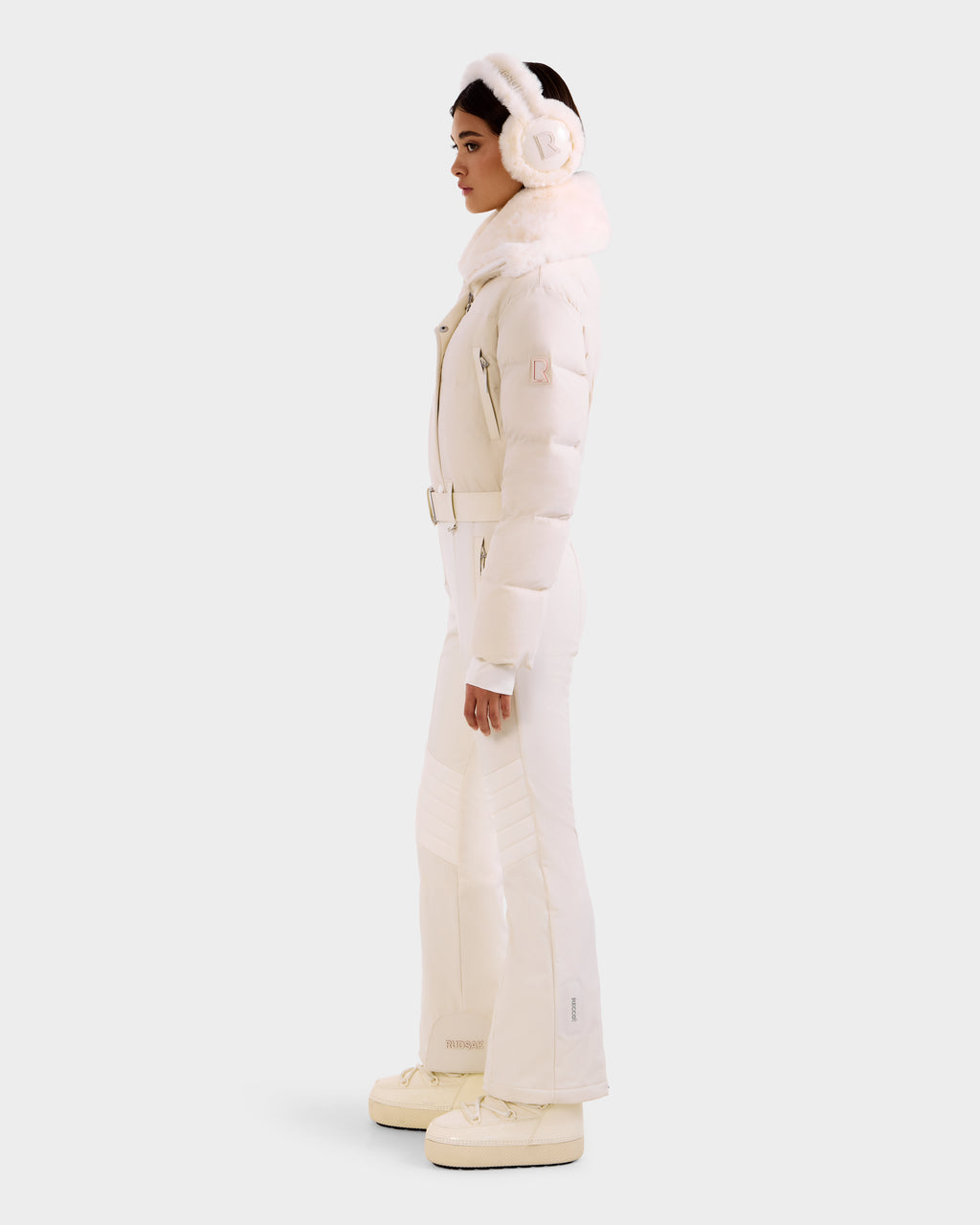 A woman in profile models Rudsak's LOVE - 8124724 Vanilla: an all-white belted puffer jacket, paired with flared pants, platform boots, and earmuffs, set against a plain light background.