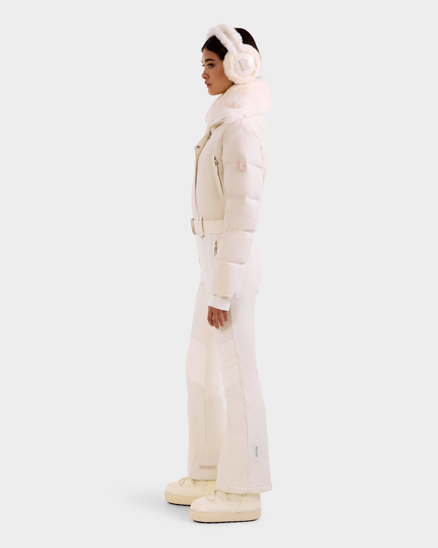 A woman in profile models Rudsak's LOVE - 8124724 Vanilla: an all-white belted puffer jacket, paired with flared pants, platform boots, and earmuffs, set against a plain light background.