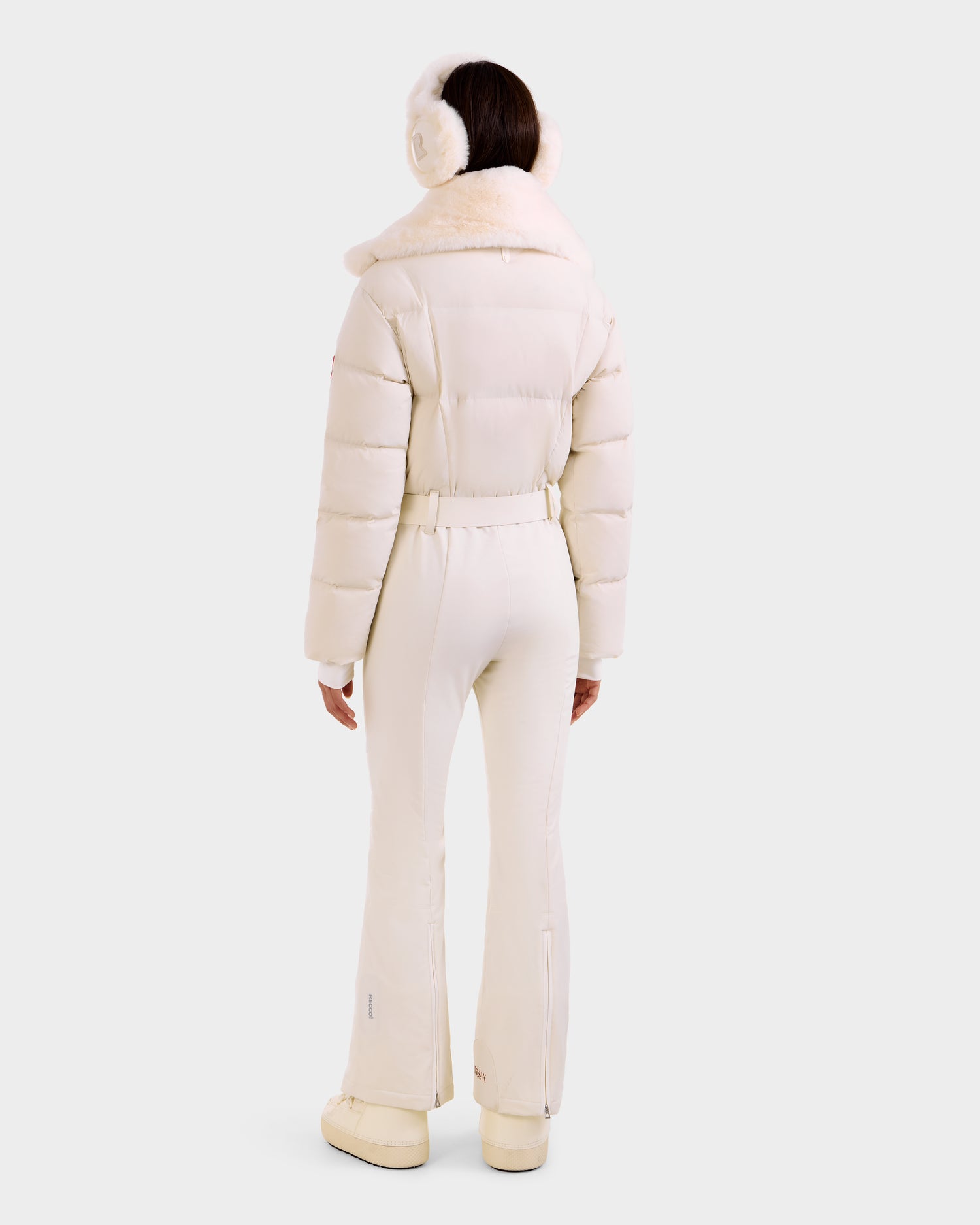 A person faces away, wearing the Rudsak LOVE - 8124724 Vanilla women's ski suit with a fur collar, white earmuffs, and boots, set against a plain light background.