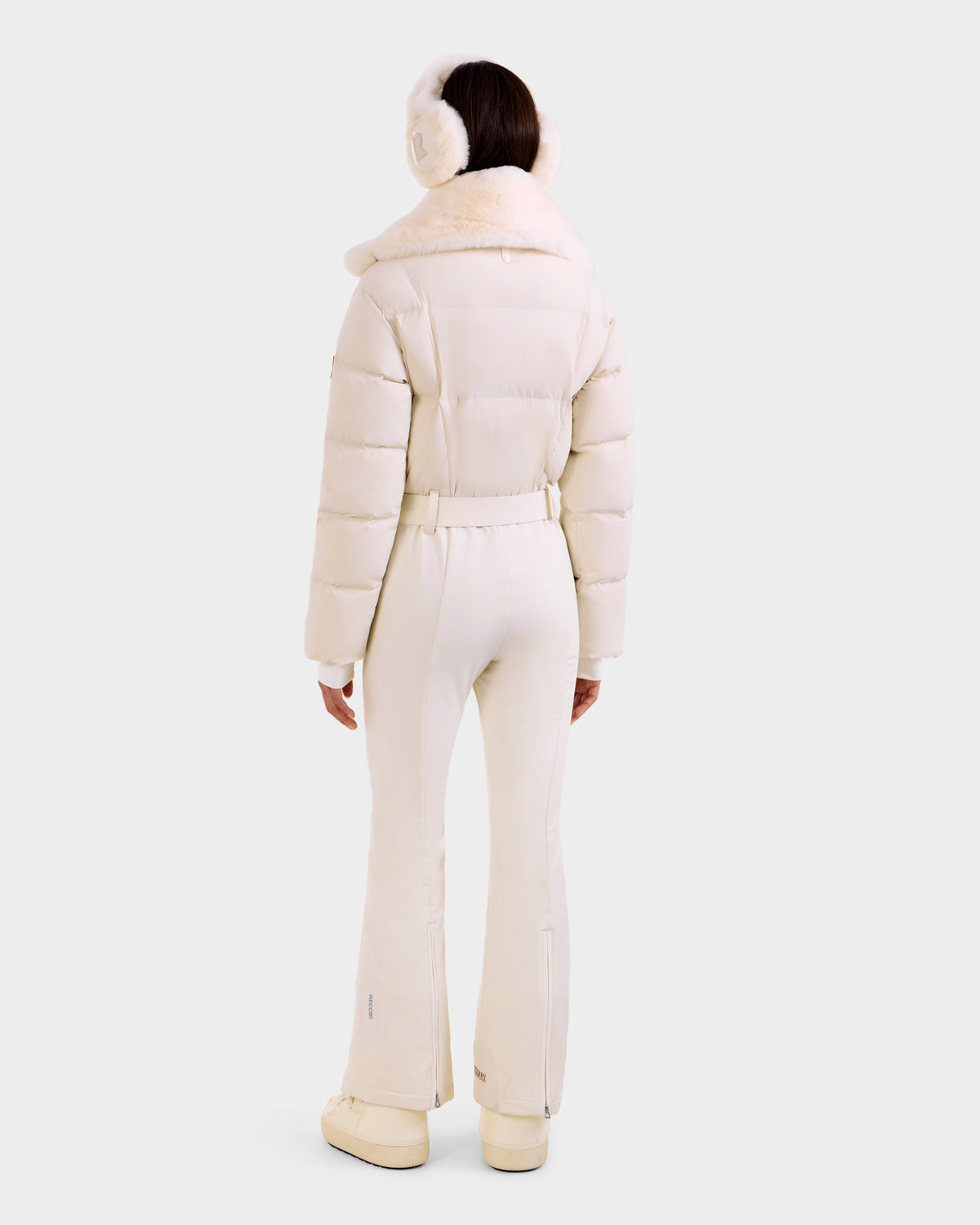 A person faces away, wearing the Rudsak LOVE - 8124724 Vanilla women's ski suit with a fur collar, white earmuffs, and boots, set against a plain light background.