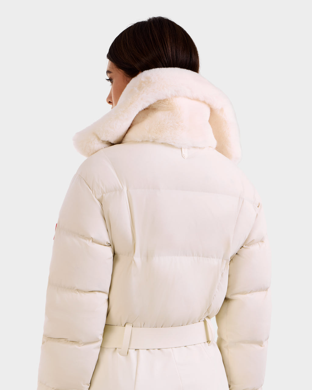 A woman with dark brown hair wears the Rudsak LOVE - 8124724 Vanilla puffer coat, featuring a wide plush fur collar and belt. The stylish, warm women’s coat stands out against a plain light background.