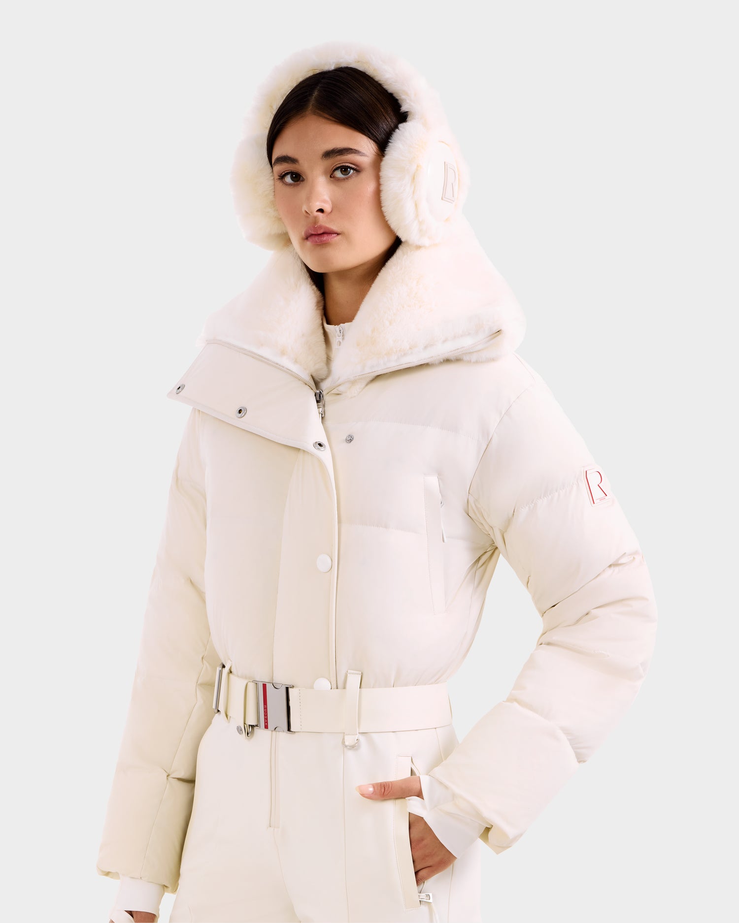 A woman poses against a plain light background wearing the Rudsak LOVE - 8124724 Vanilla ski suit, featuring a wide collar, matching earmuffs, and high-waisted belted pants, with one hand in her pocket.
