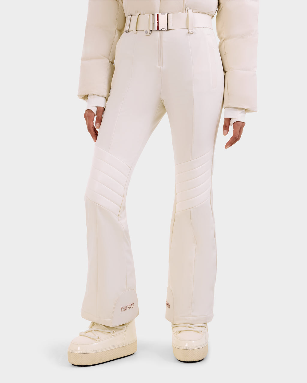 A person wearing the Rudsak LOVE - 8124724 Vanilla women's ski suit with a matching belt, quilted knees, a puffy jacket, and platform snow boots featuring RECCO rescue technology, set against a white background.