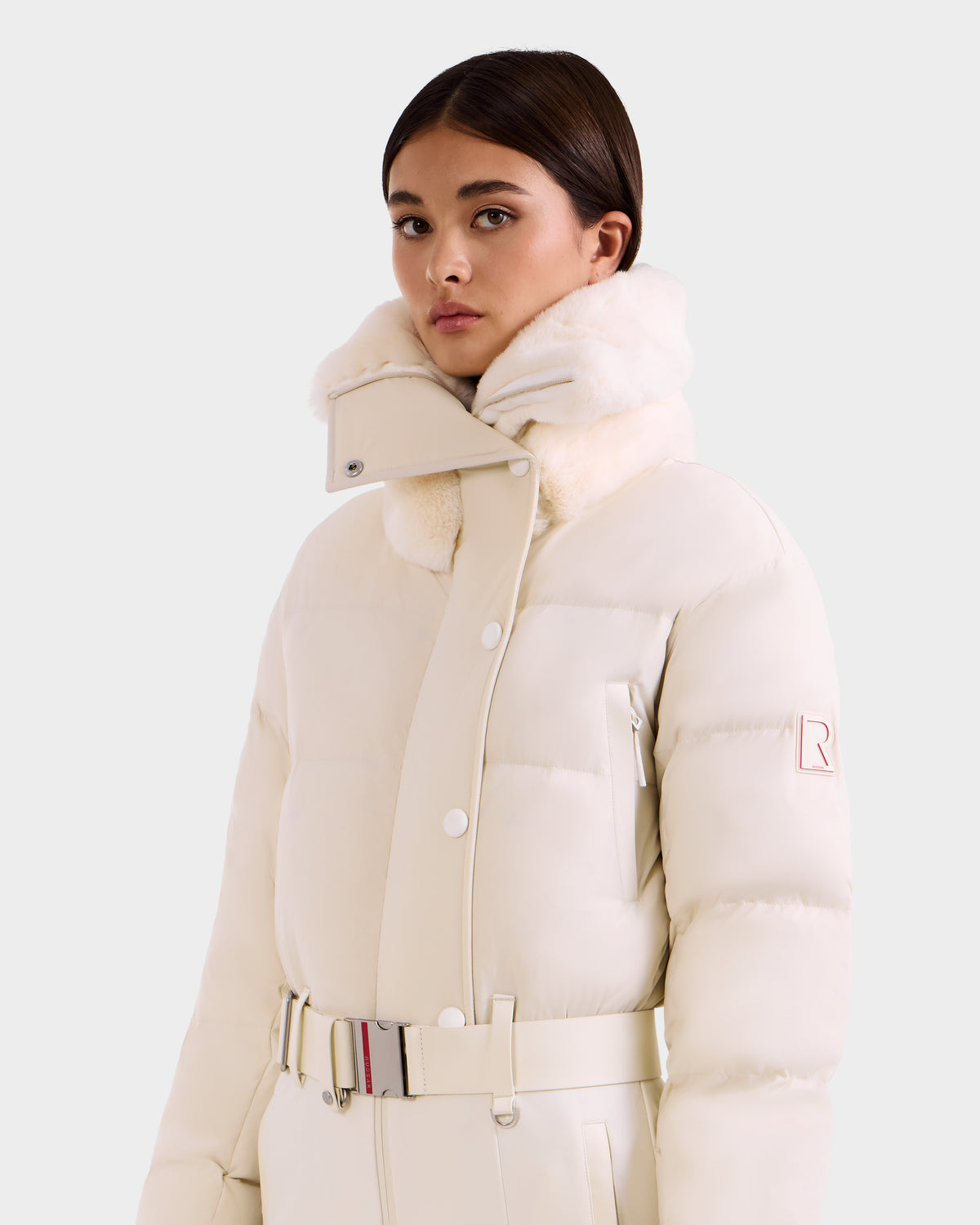 A young woman with straight dark hair wears the Rudsak LOVE - 8124724 Vanilla high-collared, belted off-white puffer jacket with RECCO rescue technology and a sleeve patch, standing against a plain light background.
