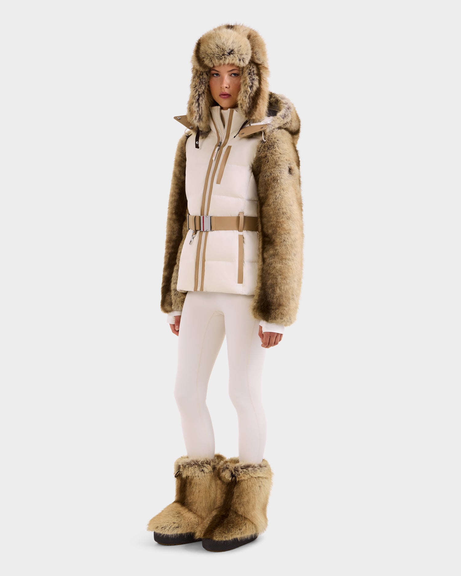 A person wears the Rudsak JOANNA F - 8125593 Mocha cream and tan faux-fur ski jacket with RECCO rescue tech, paired with a beige belt and white leggings, against a plain white background.