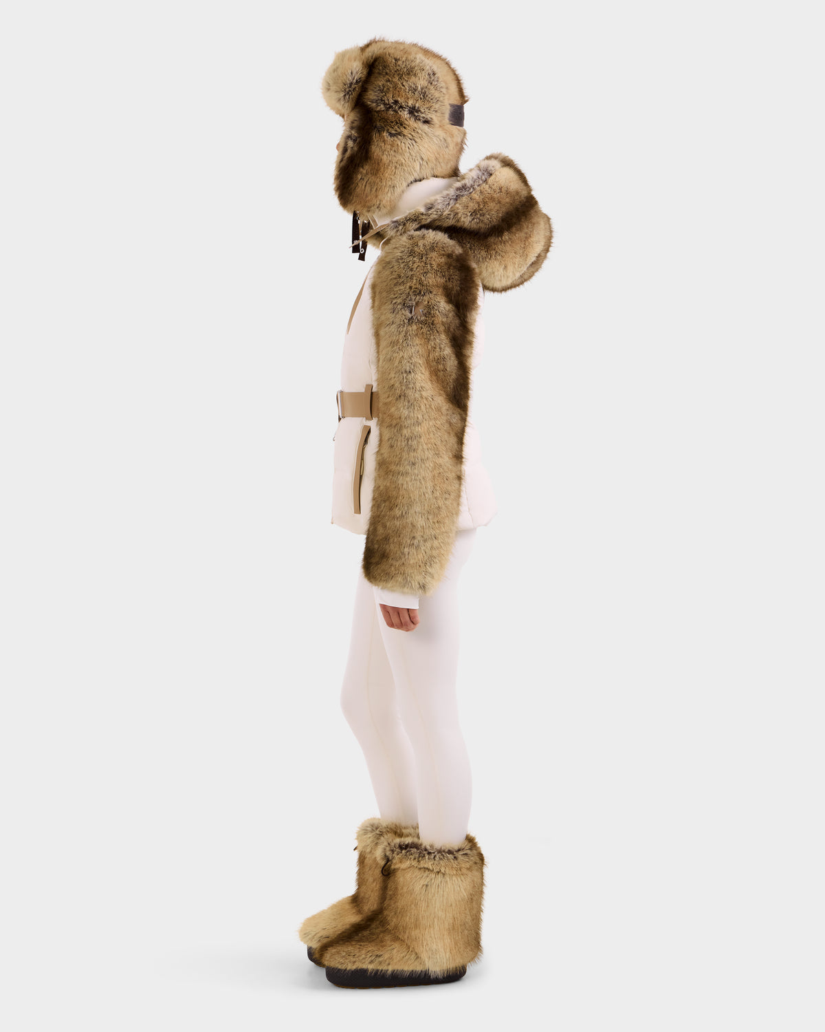 A person stands in profile in the Rudsak JOANNA F - 8125593 Mocha women’s down ski jacket with faux fur trim and a hood, styled with mittens, tall boots, fitted pants, and a belt, all set against a plain white background.