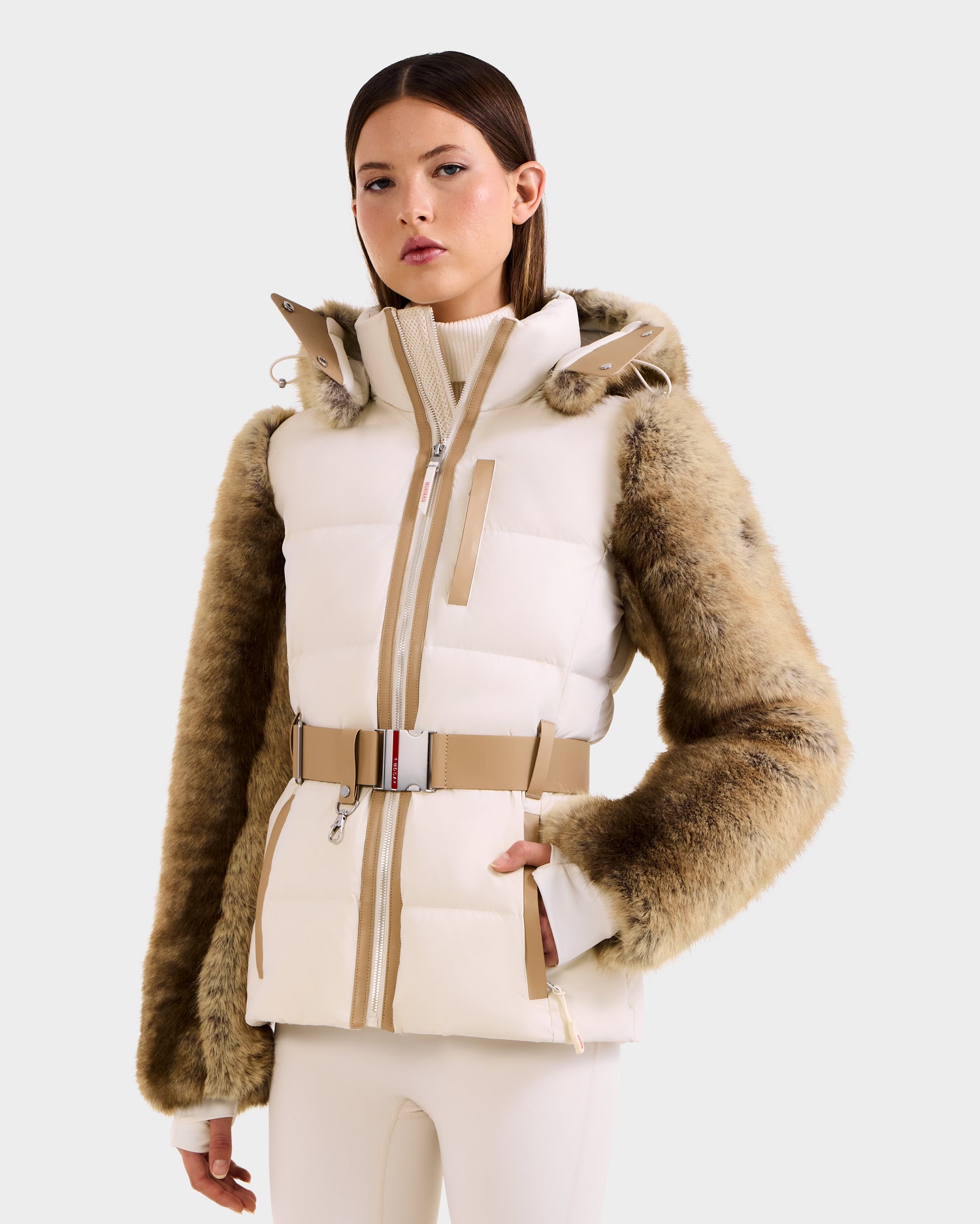 A woman models the Rudsak JOANNA F - 8125593 Mocha faux-fur ski jacket with beige fur sleeves, a belted waist, and high collar, styled with white pants against a plain light background.