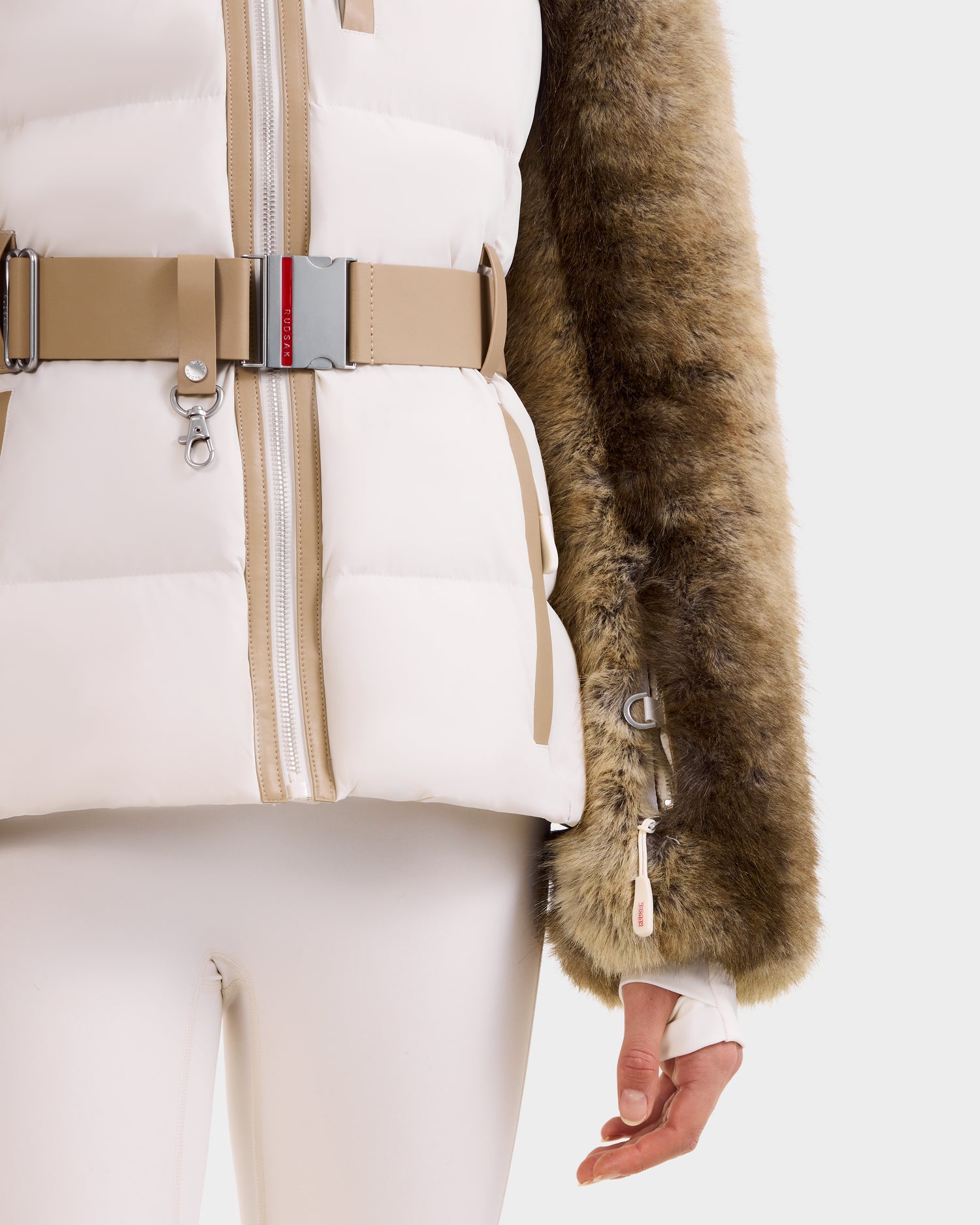 A person models the Rudsak JOANNA F - 8125593 Mocha women’s down ski jacket with a white quilted vest, beige accents, a belt, and a brown faux fur sleeve, paired with white pants. Only the lower torso and right arm show against a plain light background.