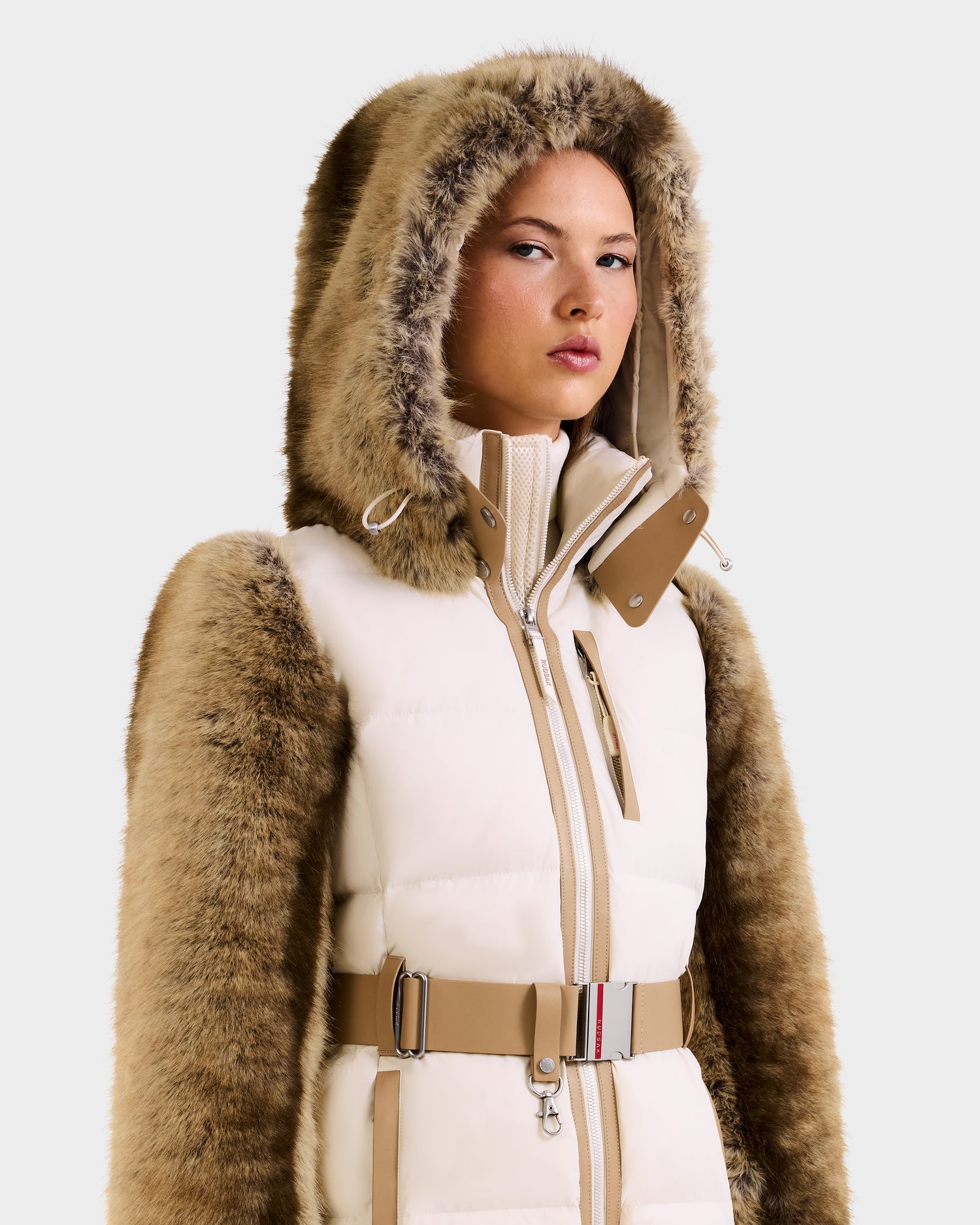 A person models the Rudsak JOANNA F - 8125593 Mocha faux-fur ski jacket, featuring a high collar, beige belt, and zipper details, against a light plain background.