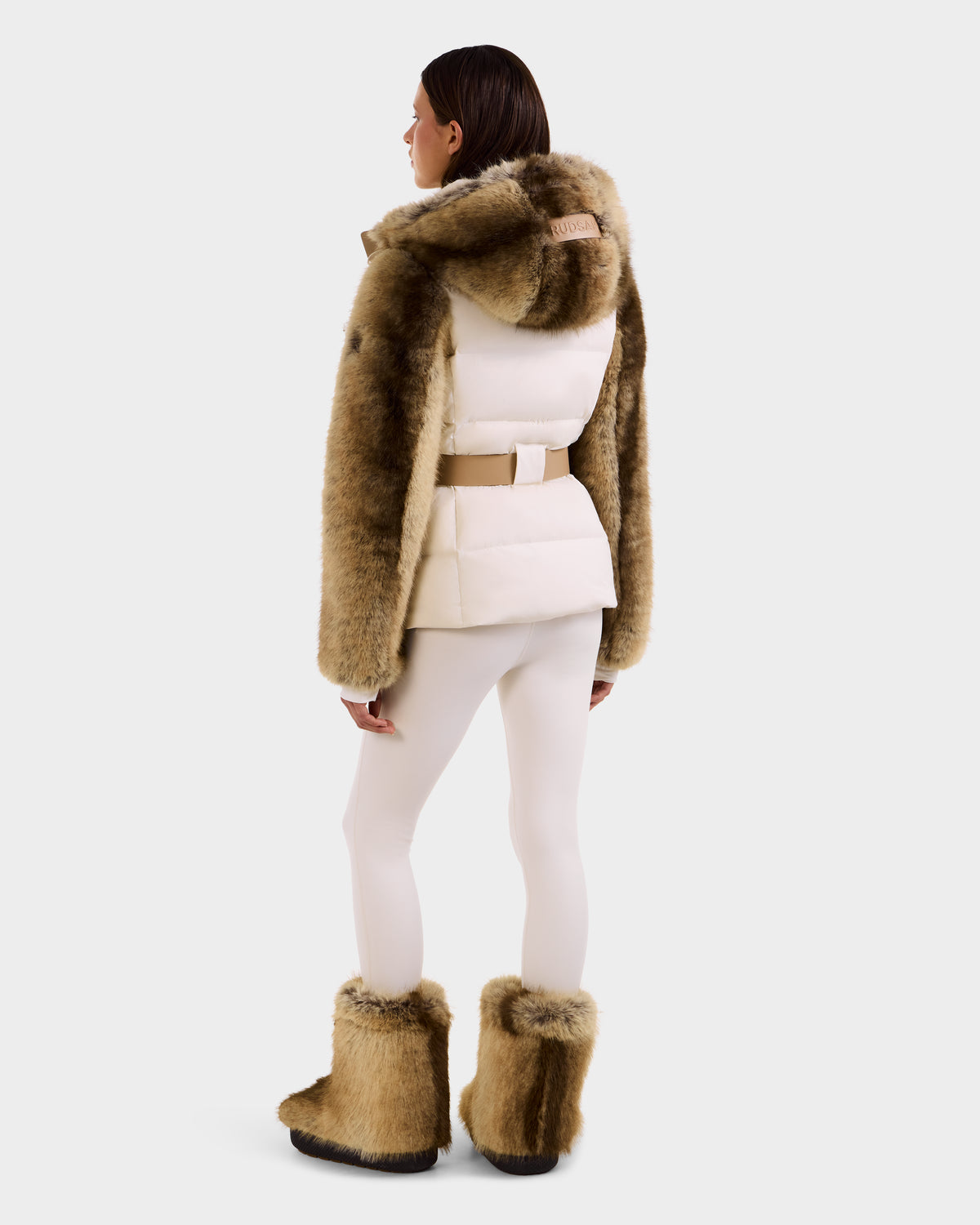 A woman faces away, wearing the Rudsak JOANNA F - 8125593 Mocha faux-fur ski jacket with hood, matching boots, beige belt, and white leggings against a plain background.