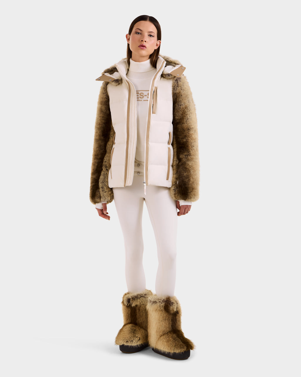 A woman stands against a white background wearing the Rudsak JOANNA F - 8125593 Mocha faux-fur ski jacket with brown fur sleeves, matching boots, white leggings, and a turtleneck top, facing forward with a neutral expression.