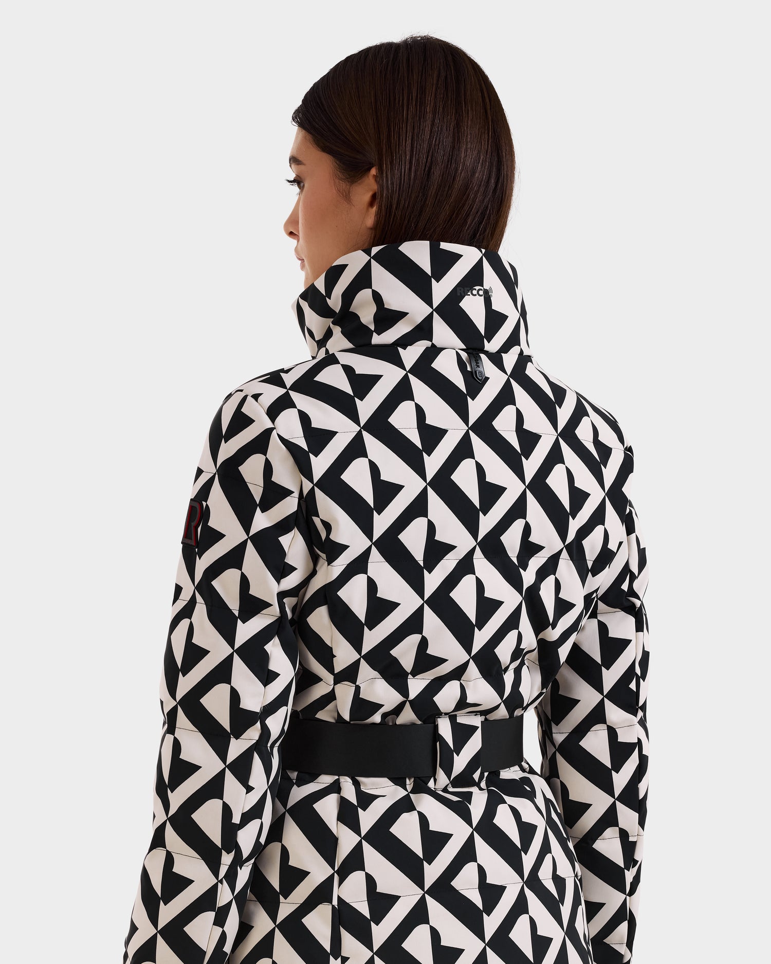 A woman with straight brown hair wears the Rudsak JOANNA P - 8125623 Vanilla Monogram coat, featuring a black-and-white geometric pattern, high collar, belted waist, and RECCO® rescue technology, shown from the back against a light background.
