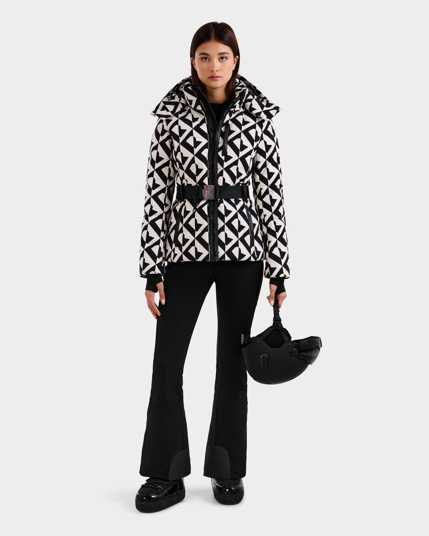 A woman with straight dark hair and a neutral expression stands against a white background, holding a black ski helmet and wearing black flared pants, boots, and the Rudsak JOANNA P - 8125623 Vanilla Monogram women’s down ski jacket with removable hood.