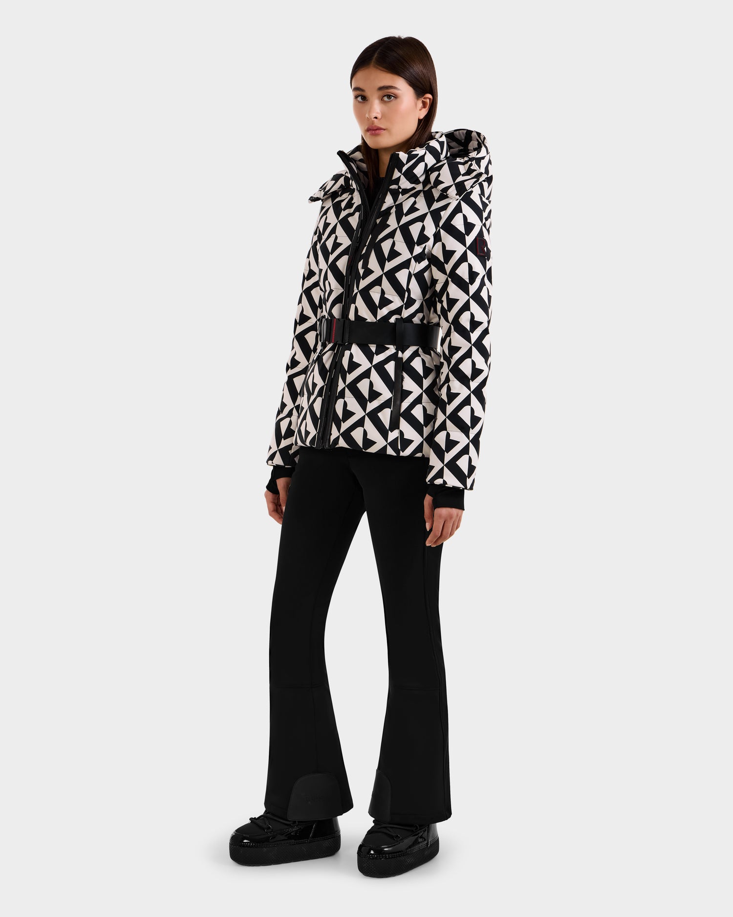 A woman wears the Rudsak JOANNA P - 8125623 Vanilla Monogram down ski jacket with removable hood and belt, paired with black flared pants and shoes, standing against a plain light background.