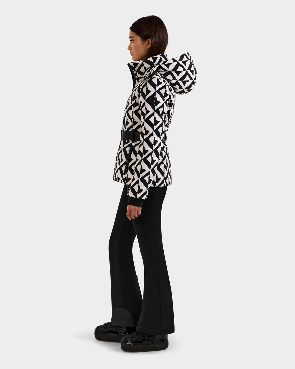 A woman stands in profile wearing the Rudsak JOANNA P - 8125623 Vanilla Monogram women’s down ski jacket with a removable hood, paired with black flared pants and black shoes against a plain light background.