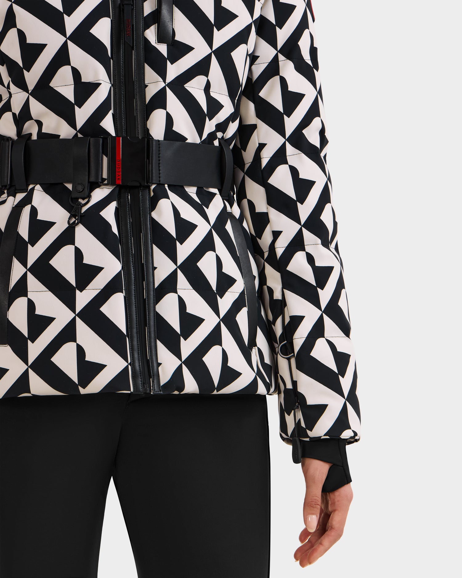 A person wears the Rudsak JOANNA P - 8125623 Vanilla Monogram women’s ski jacket, featuring a black and white pattern with heart and triangle shapes, a black belt, and black pants. Only the lower torso and one hand are shown against a plain background.