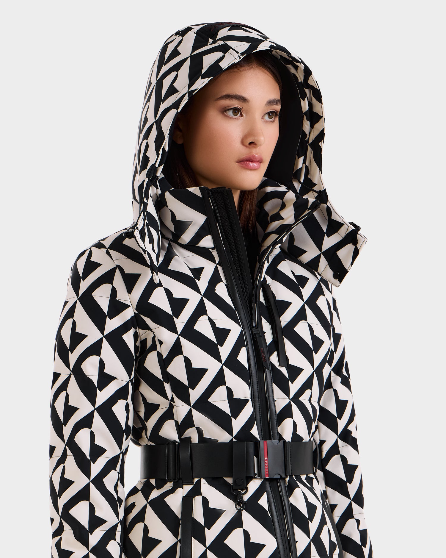 A woman wearing the Rudsak JOANNA P - 8125623 Vanilla Monogram down ski jacket with a hood and monogram pattern, belted at the waist, looks slightly to the side against a plain light background.