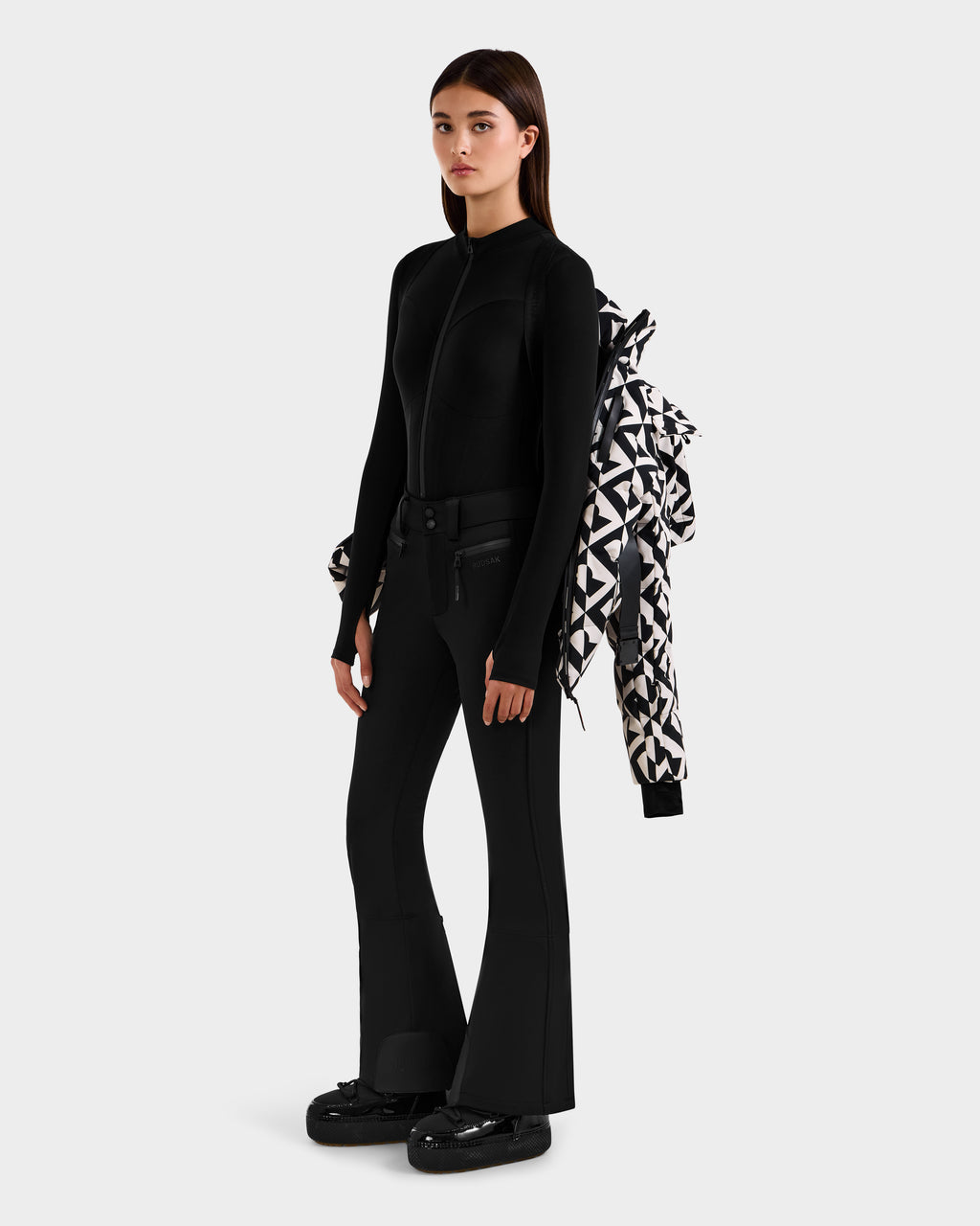 A woman stands against a plain background wearing a sleek black jacket and flared pants, holding the Rudsak JOANNA P - 8125623 Vanilla Monogram down ski jacket over her arm and wearing chunky black boots.