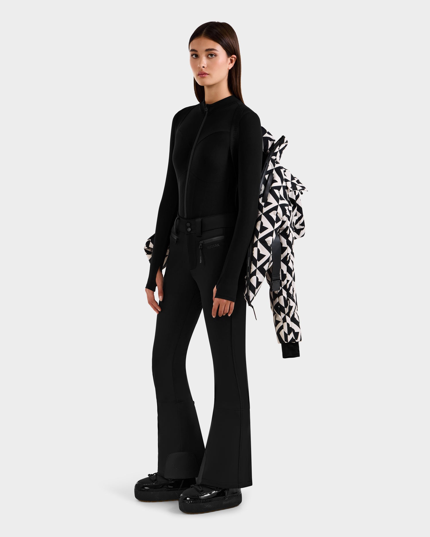 A woman stands against a plain background wearing a sleek black jacket and flared pants, holding the Rudsak JOANNA P - 8125623 Vanilla Monogram down ski jacket over her arm and wearing chunky black boots.