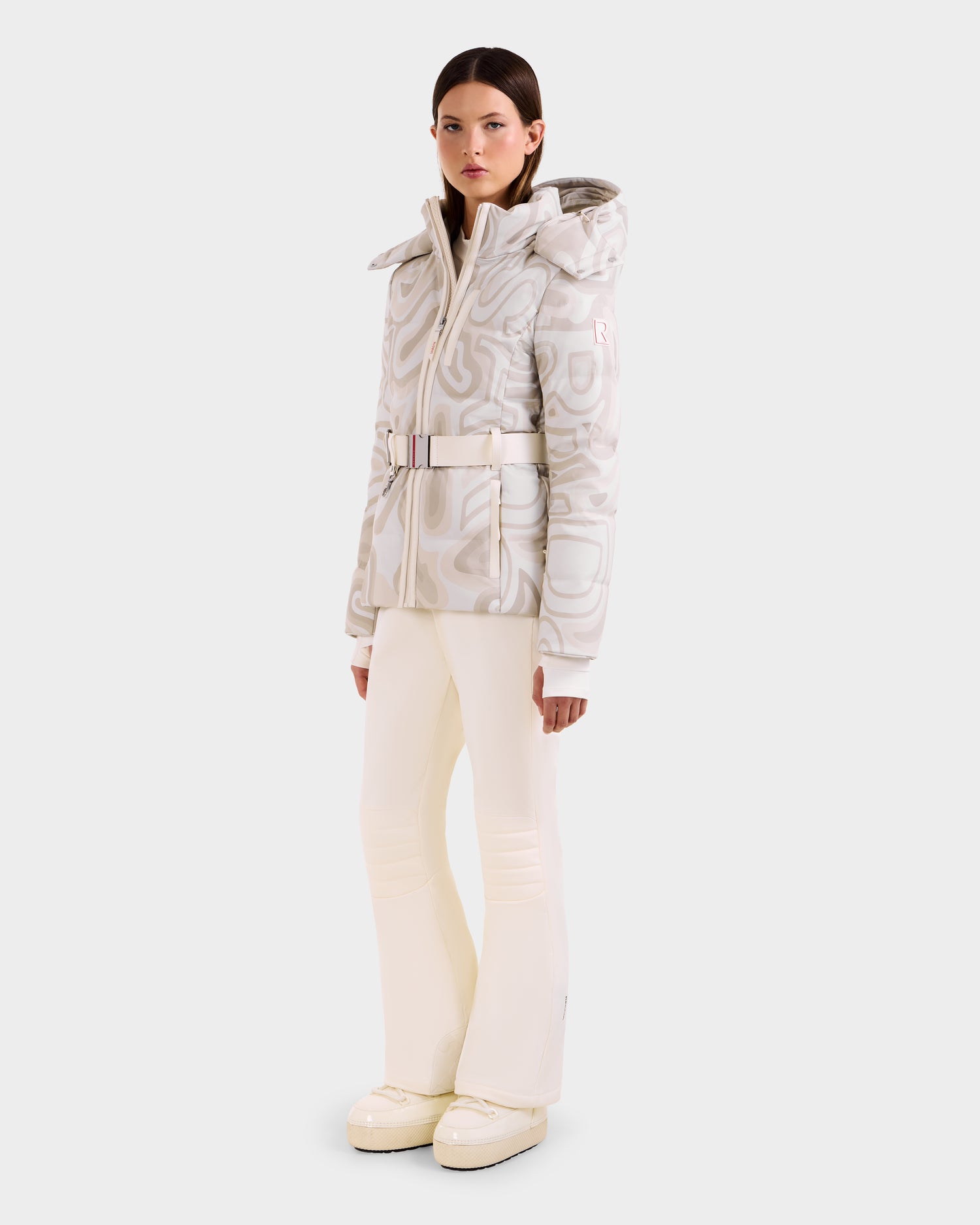 A woman with straight brown hair wears the Rudsak JOANNA P - 8125623 Vanilla Deja vu women’s down ski jacket and matching flared pants, styled with platform boots, posing against a plain light background.