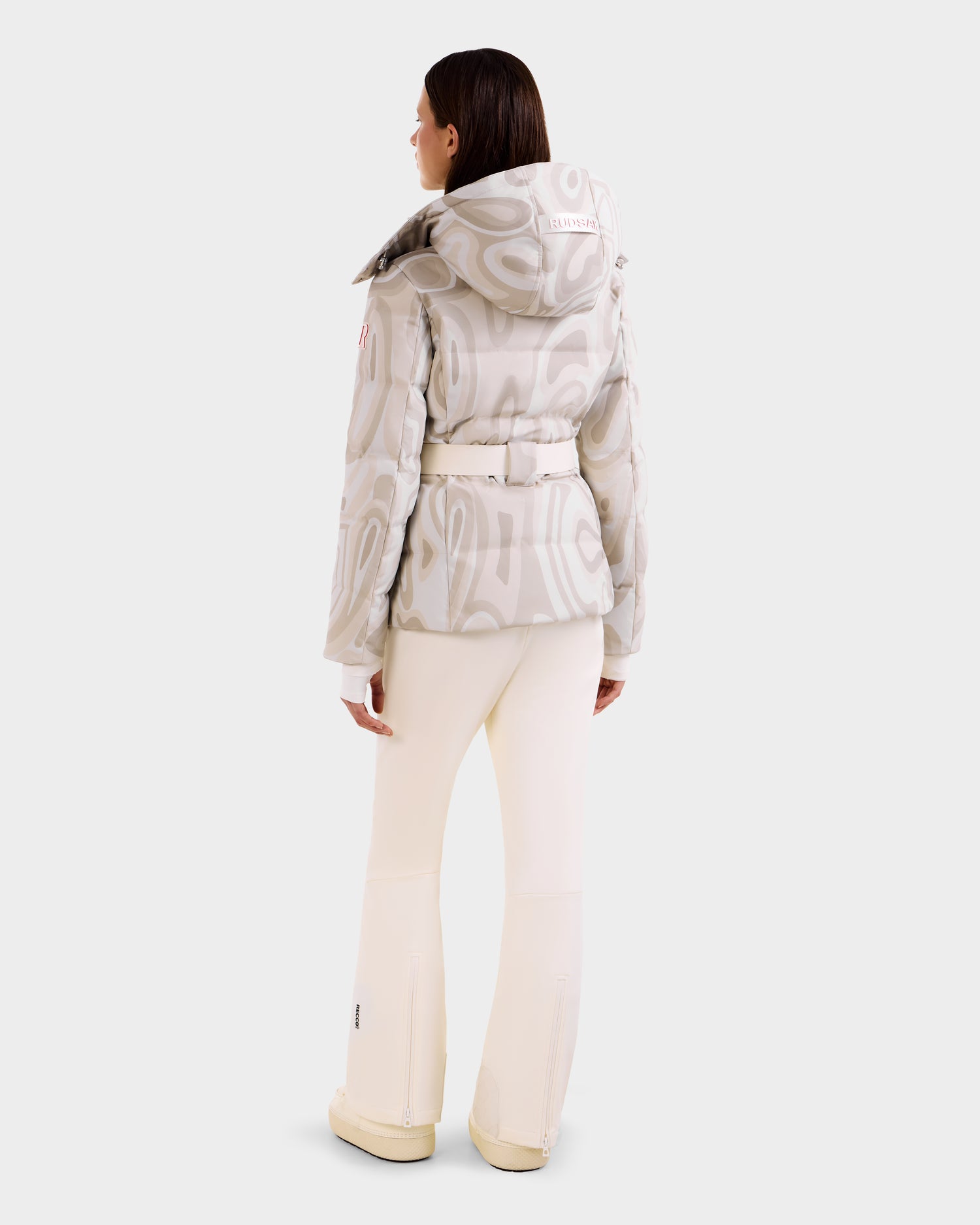 A woman faces away, wearing the Rudsak JOANNA P - 8125623 Vanilla Deja vu down ski jacket with a removable hood, paired with matching cream pants and shoes, set against a plain white background.