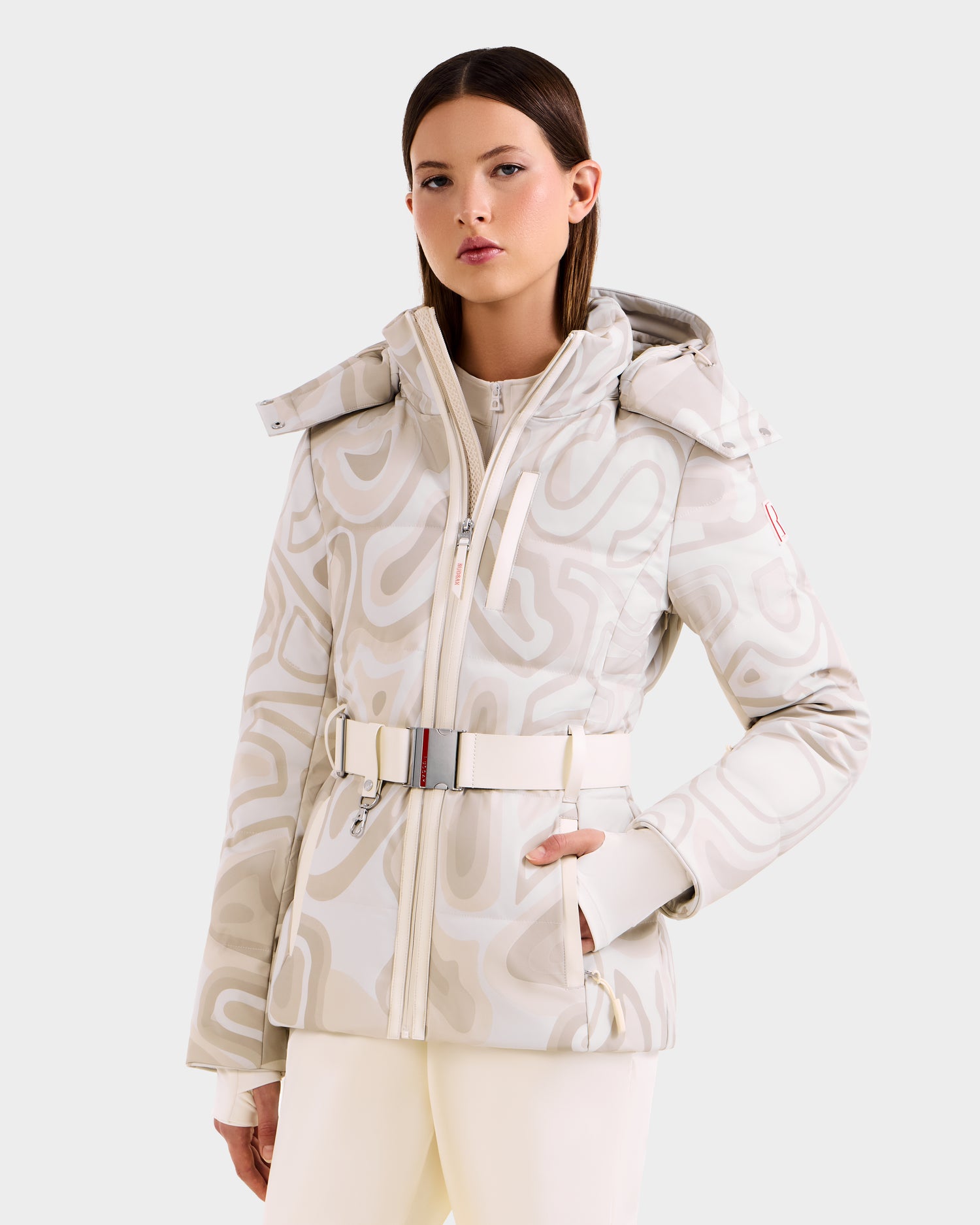 A woman with straight brown hair and a neutral expression models the Rudsak JOANNA P - 8125623 Vanilla Deja vu women’s down ski jacket, featuring a beige and white pattern, belted waist, and removable hood against a simple light background.