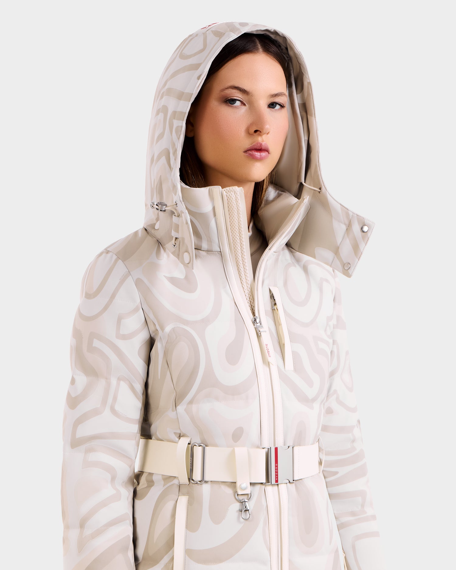 A woman stands confidently against a plain background, wearing the Rudsak JOANNA P - 8125623 Vanilla Deja vu women's down ski jacket with a removable hood and wide belt, featuring a stylish light-colored pattern.