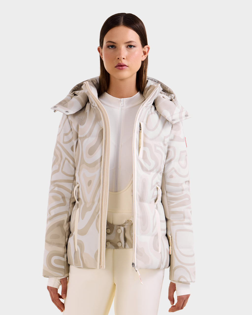 A woman wears the Rudsak JOANNA P - 8125623 Vanilla Deja vu women’s down ski jacket, white and beige patterned with a high collar, paired with matching light pants, standing against a plain light background, facing forward neutrally.
