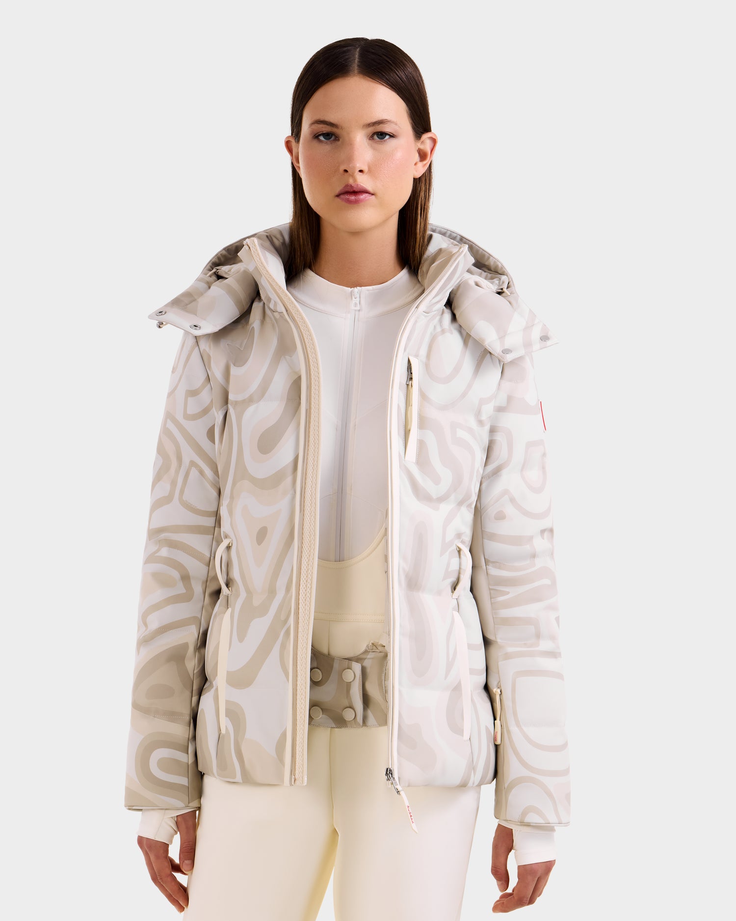 A woman wears the Rudsak JOANNA P - 8125623 Vanilla Deja vu women’s down ski jacket, white and beige patterned with a high collar, paired with matching light pants, standing against a plain light background, facing forward neutrally.