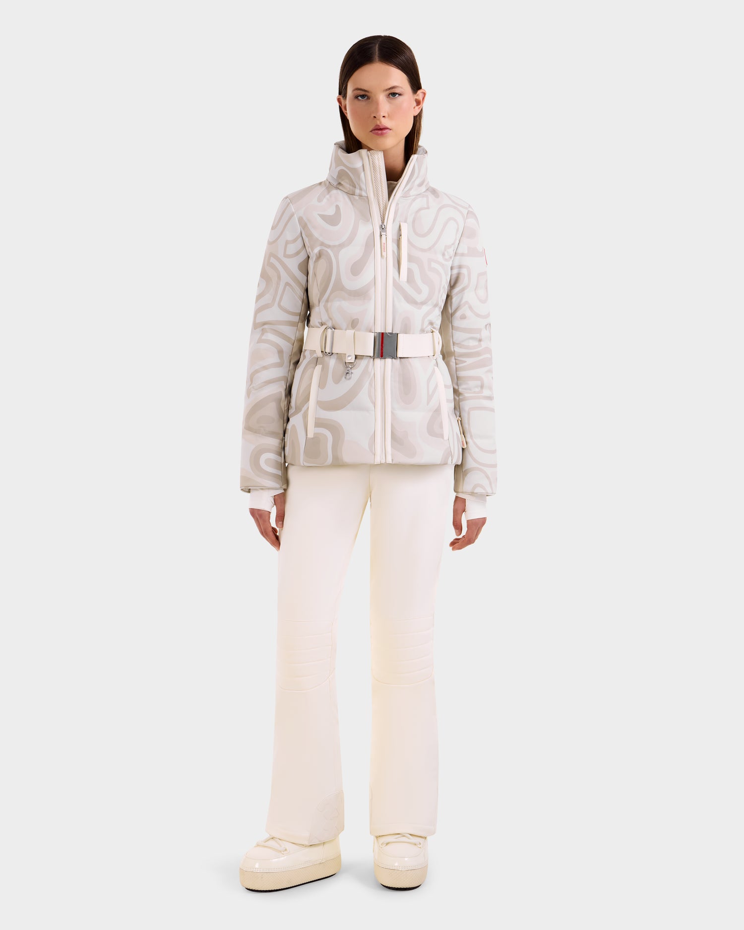 A woman faces forward, wearing the Rudsak JOANNA P - 8125623 Vanilla Deja vu patterned light gray and white women’s down ski jacket with a belted waist, paired with white pants and platform boots against a plain light background.