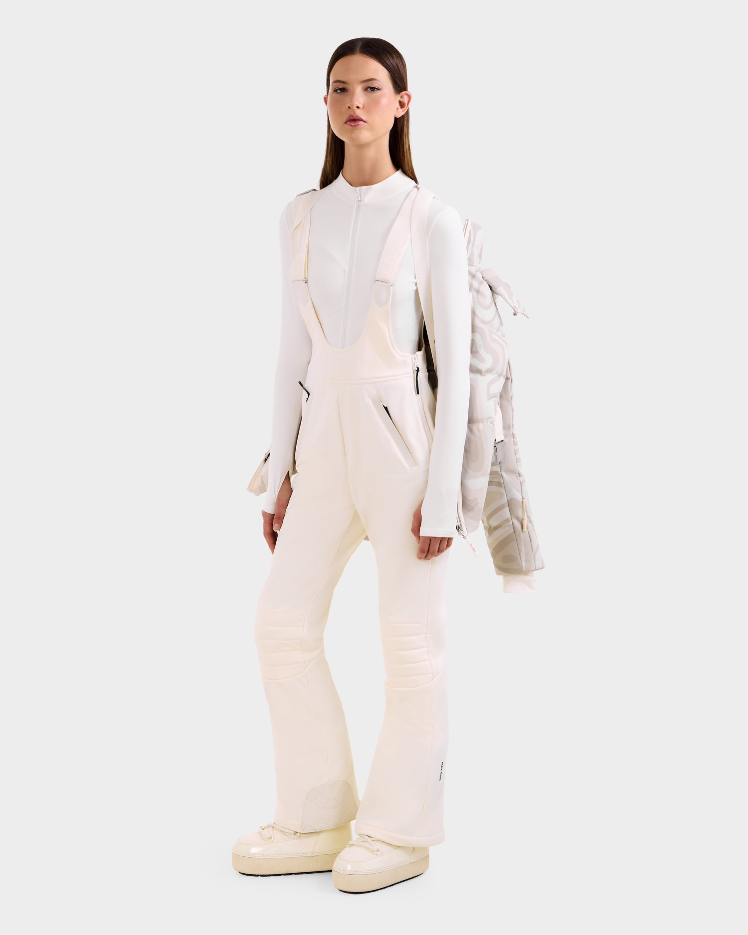 A woman faces forward in a white fitted long-sleeve top, overalls with black zippers, chunky white boots, and drapes the Rudsak JOANNA P - 8125623 Vanilla Deja vu women’s down ski jacket with removable hood over one shoulder. The background is light gray.