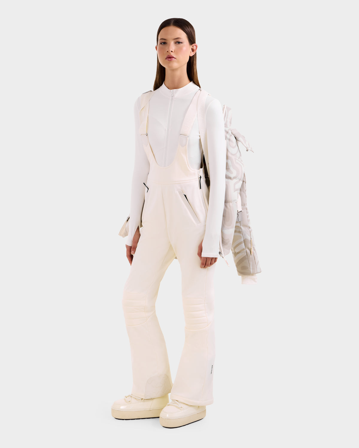A woman faces forward in a white fitted long-sleeve top, overalls with black zippers, chunky white boots, and drapes the Rudsak JOANNA P - 8125623 Vanilla Deja vu women’s down ski jacket with removable hood over one shoulder. The background is light gray.