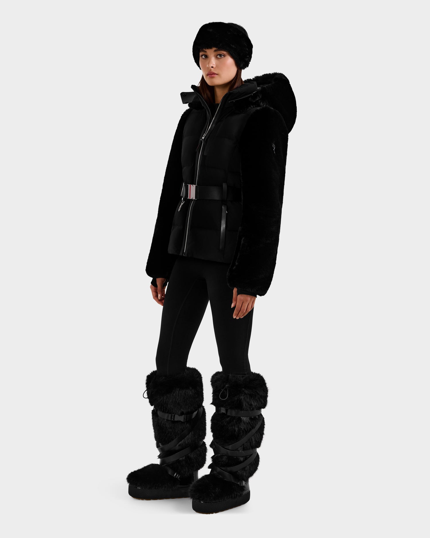 A person in an all-black winter look wears the Rudsak JOANNA F - 8125593 Black women’s down ski jacket with faux fur, paired with fitted pants and tall, furry snow boots, standing against a plain white background.