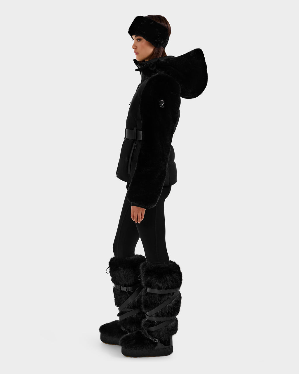 A woman in profile models the Rudsak JOANNA F - 8125593 Black faux-fur ski jacket with a hood, styled with black leggings, fur boots, and a matching headband against a plain light gray background.