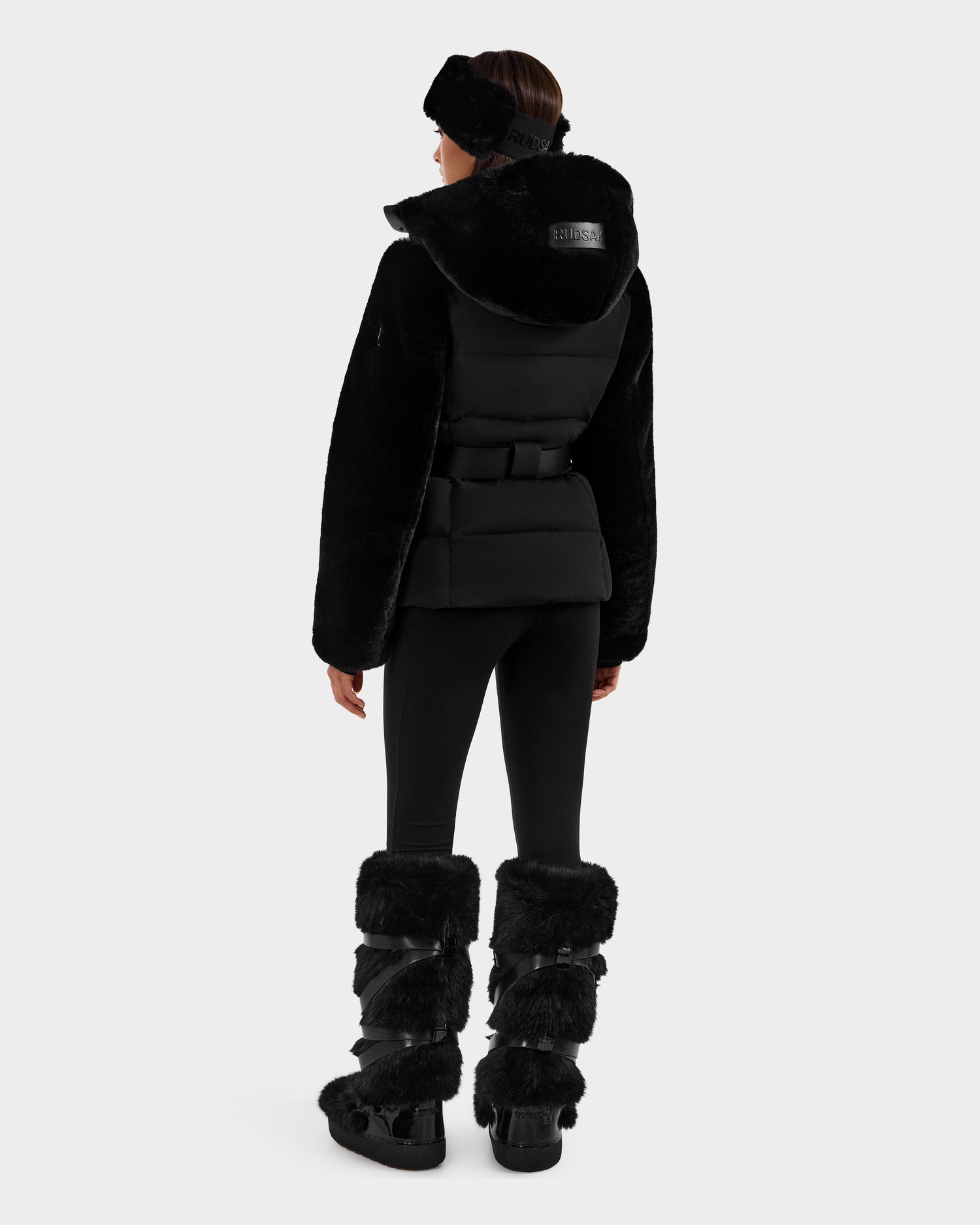 A person faces away wearing the Rudsak JOANNA F - 8125593 Black faux-fur ski jacket with a fur-trimmed hood, black leggings, matching fur boots, and a headband. The stylish winter look is set against a plain white background.